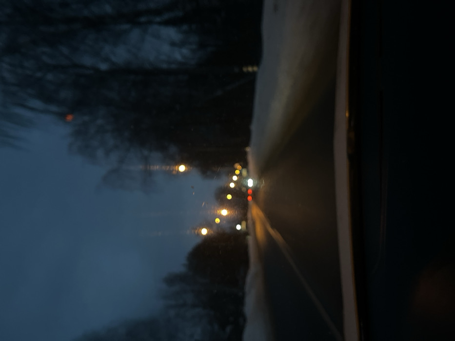 The image depicts a blurred, forward-facing view from inside a vehicle traveling on a two-lane asphalt road at dusk or night, located in Maksimkovo, Russia. Snow covers the verges on both sides of the road, which is flanked by bare trees. Multiple yellow-orange streetlights illuminate the scene, casting reflections on the wet or icy road surface. Two vehicles are visible in the distance, one with red taillights and another with white headlights. The vehicle's dashboard is visible at the bottom of the frame. No people or specific interactions are discernible. The image quality is out of focus, with some streaks or smudges visible on what appears to be the windshield.