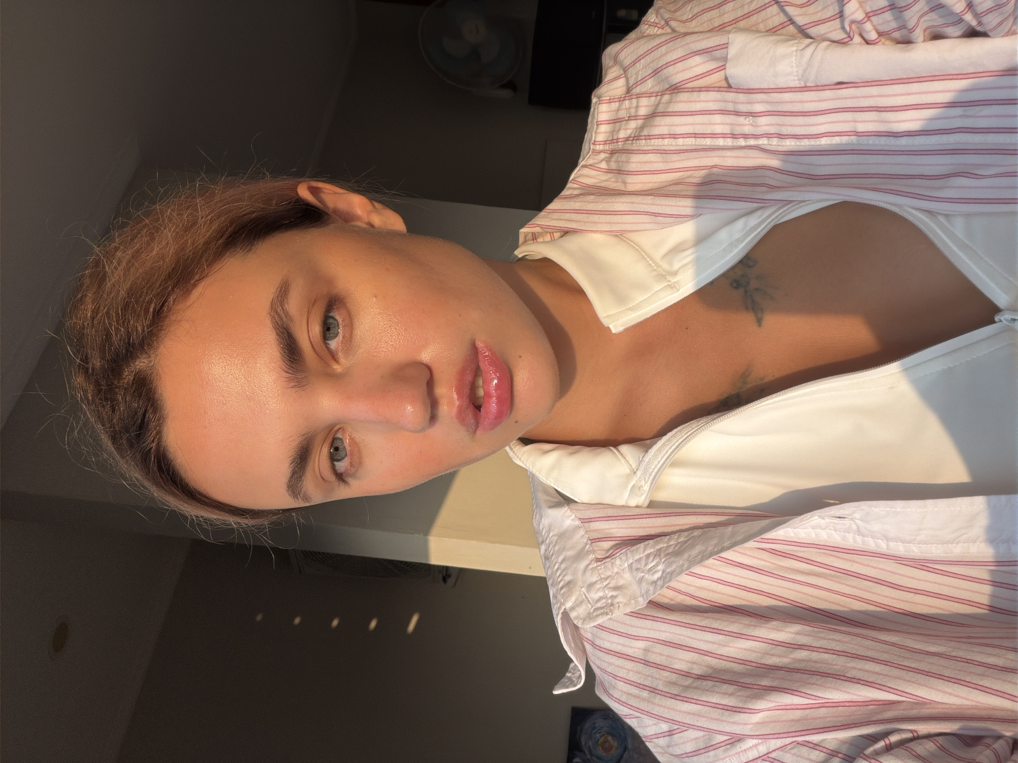 A close-up, head-and-shoulders portrait shows a young Caucasian woman with light brown hair pulled back from her face. Her eyes are light blue, and her lips are full and glossy. She is wearing a white collared shirt with thin pink stripes and an open white zipper top underneath, revealing a portion of her chest with a dark tattoo. The lighting suggests it is late afternoon, with warm sunlight casting shadows on the wall behind her. The background is slightly out of focus, showing a dark interior with some light streaks on the wall, possibly from blinds or a window. To the right, a part of a fan is visible. The overall setting appears to be an indoor space, possibly a home or hotel room, in North Kuta, Indonesia.