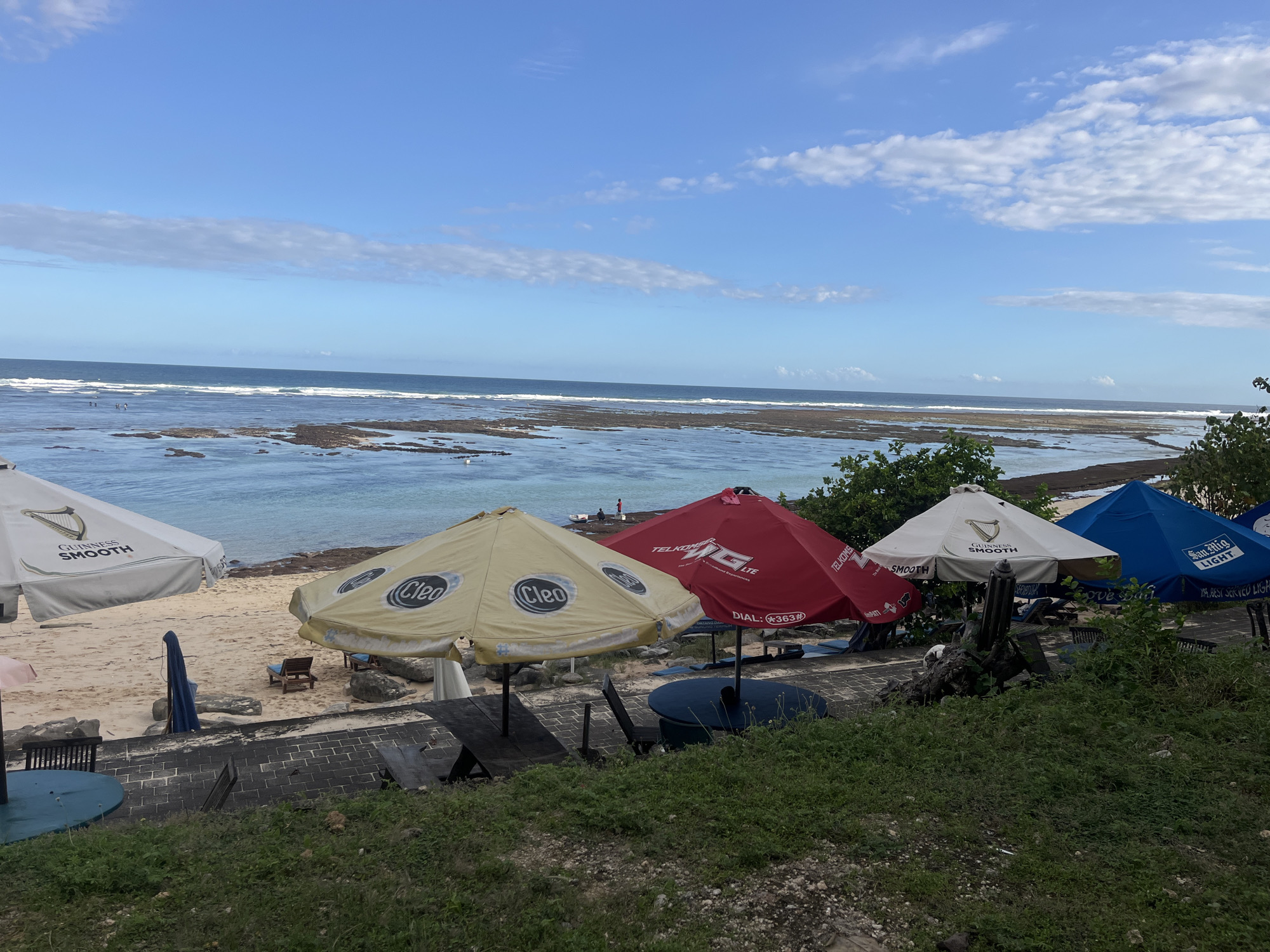 The image depicts a coastal scene in Kuta Selatan, Indonesia. The foreground is dominated by several beach umbrellas advertising different brands: "Guinness Smooth", "Cleo", "Telkomsel LTE", and "San Mig Light". Behind the umbrellas is a sandy beach with some sun loungers. Beyond the beach, there is a rocky tidal area with shallow water and some small waves. In the background, the sea stretches to the horizon under a blue sky with scattered clouds. Some people can be seen wading in the water and standing on the rocks. The foreground also includes some greenery, a paved area with tables and chairs, and some low-lying vegetation.