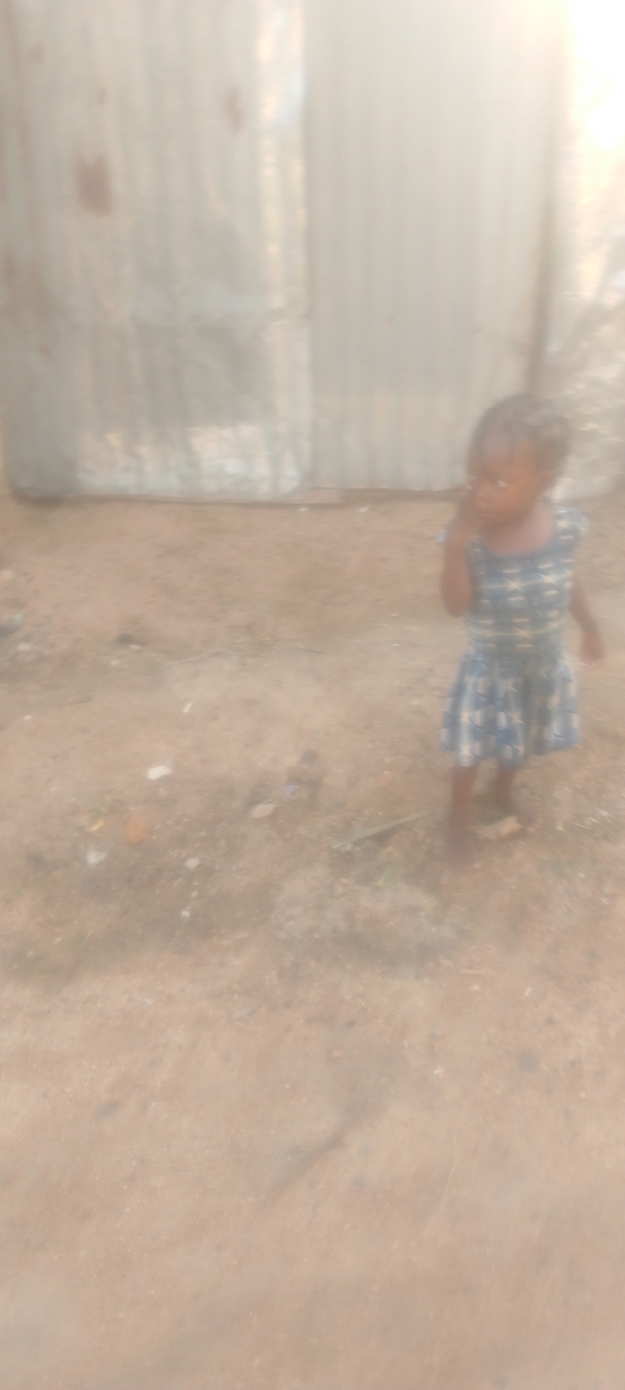 In Jalingo, Nigeria, a young child stands in front of a corrugated metal wall. The child, with dark hair braided, wears a blue and white patterned dress. The ground is dirt, with some scattered debris visible. The lighting suggests it is daytime, with a bright light source coming from the right side of the image. The overall scene appears to be an outdoor, perhaps informal, setting.