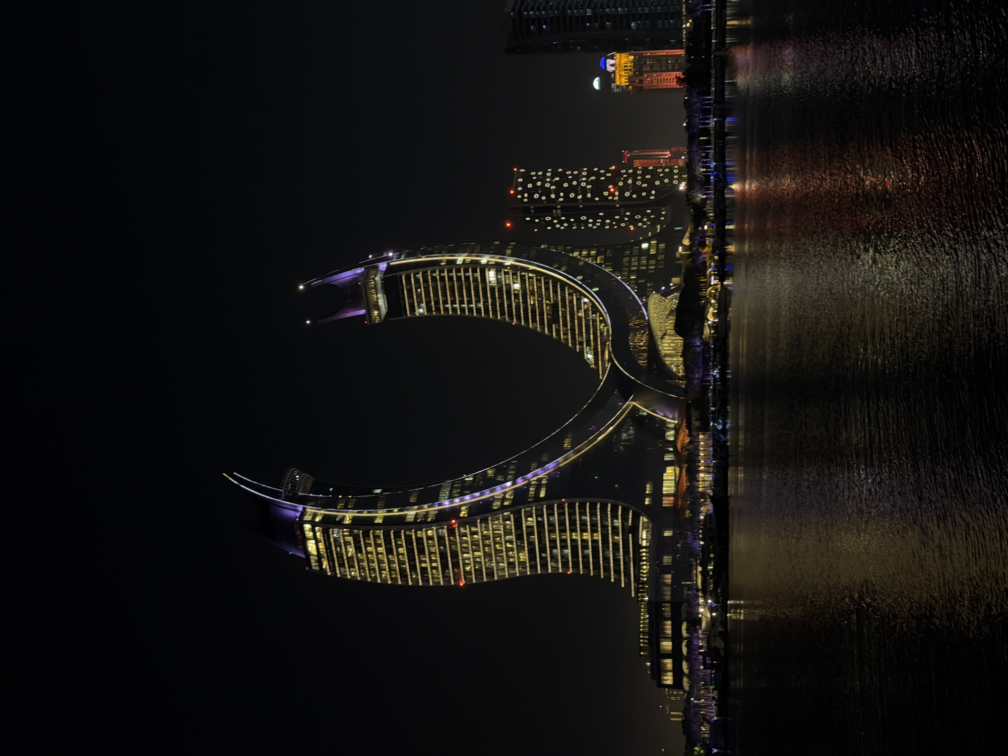 This is a nighttime, vertical, outdoor shot of a modern cityscape with a body of water in the foreground. The primary subjects are a series of illuminated buildings and their reflections in the water. The architecture is contemporary, with one prominent building featuring a distinctive crescent or C-shaped design, outlined in purple lights and filled with horizontal rows of yellow lights. Adjacent buildings also display a grid of illuminated windows. The water's surface is textured and dark, reflecting the lights of the buildings in a shimmering, elongated pattern of reds, yellows, and purples. The scene is illuminated by artificial light from the buildings, creating a sense of urban vibrancy against the dark night sky. There are no people or specific activities clearly visible, suggesting a general view of a city at night. The overall mood is one of impressive modern architecture and urban development. No text is visible in the image.