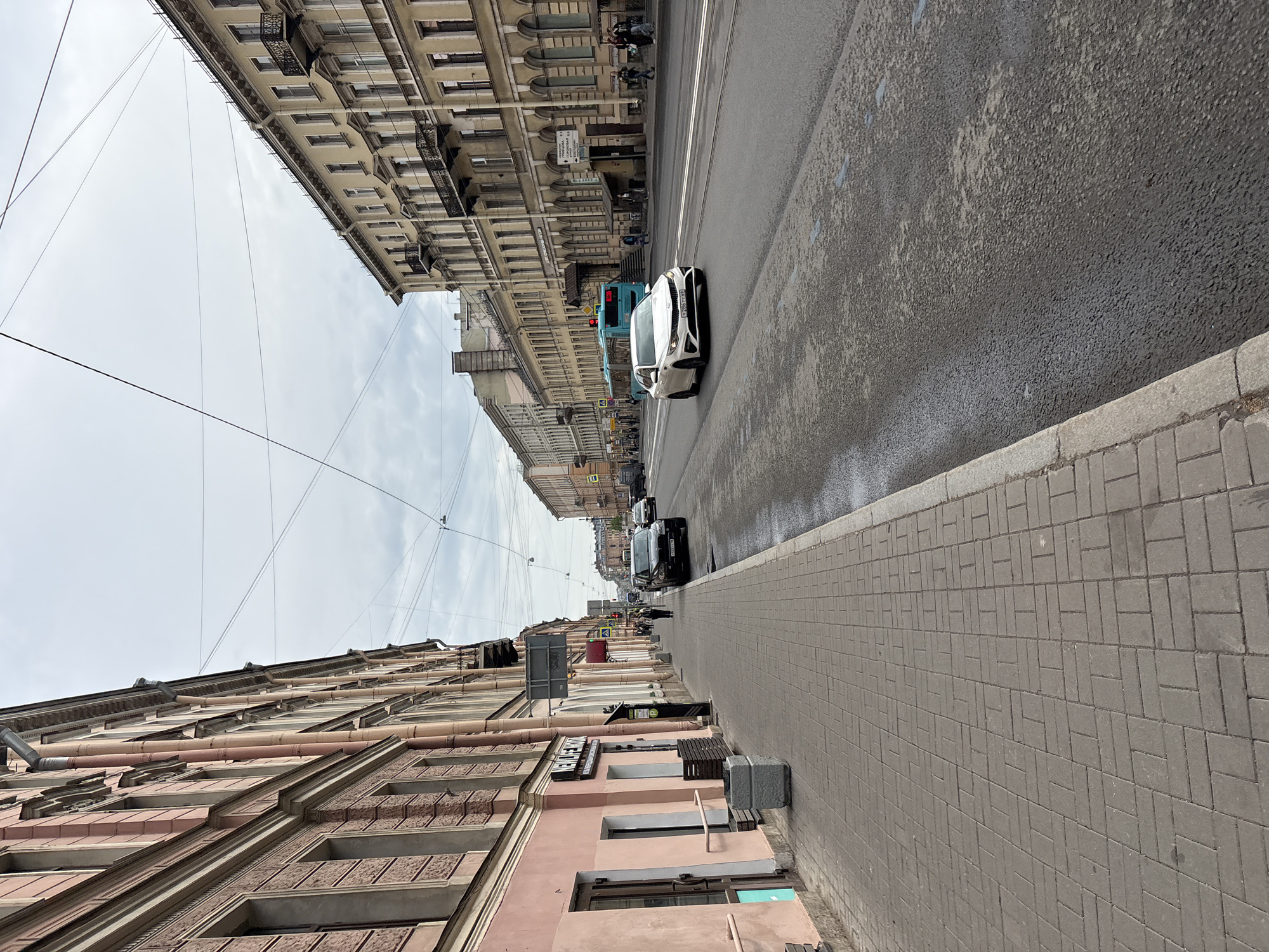 The image depicts a street in Saint Petersburg, Russia. The perspective is from a low angle, capturing the sidewalk on the right, the street in the middle, and buildings on both sides. The sky is overcast.
On the right, the sidewalk is paved with rectangular gray bricks. The buildings on the right side are several stories high, with a mix of pink and brick facades. Signage is visible on the buildings.
In the street, cars and a bus are driving. The road surface is wet and has visible tram lines.
The buildings on the left side are taller and more ornate than those on the right, with lighter-colored facades. Overhead, there are crisscrossing electrical wires.