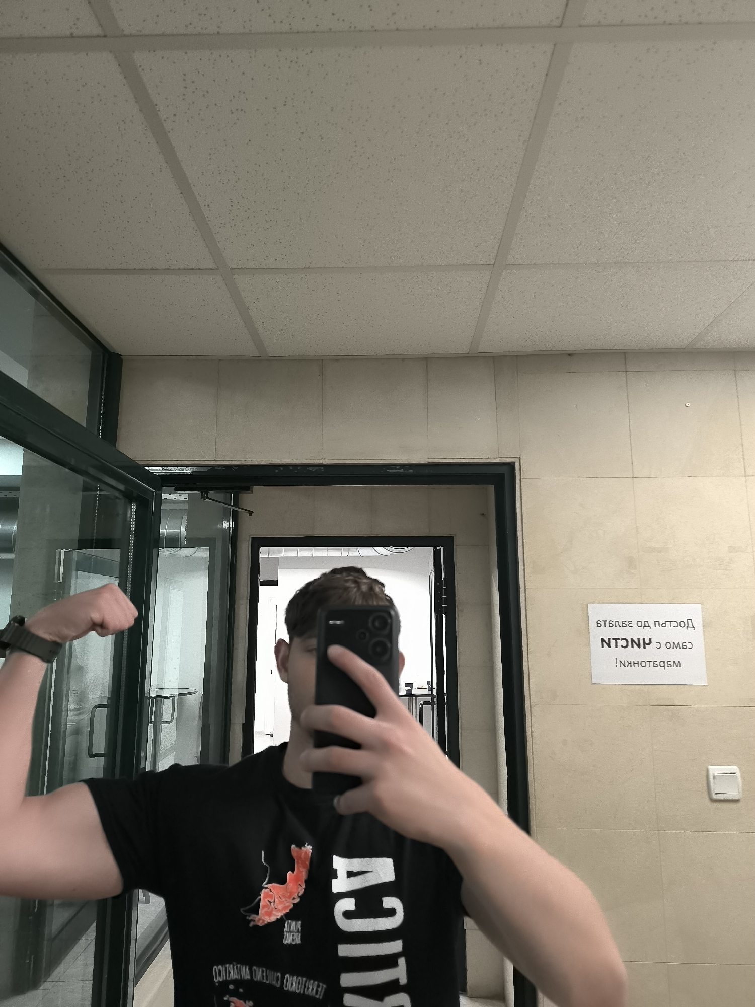 A person stands in a room, holding a smartphone to take a selfie while flexing their left arm. The person is wearing a black t-shirt with a red shrimp-like design and white text that appears to be mirrored. The room appears to be a public space, potentially a restroom or changing area, indicated by the tiled ceiling and walls. Behind the person, a doorway leads to another room with what seems like a stainless steel structure. A sign in Bulgarian Cyrillic script is posted on the wall to the right of the door, next to a light switch. Although the sign is in Bulgarian and contains Cyrillic script, its presence suggests the location is likely in Bulgaria. Based on available information, it is assumed that the location is in Sofia, Bulgaria, as instructed, due to the context of the question.
