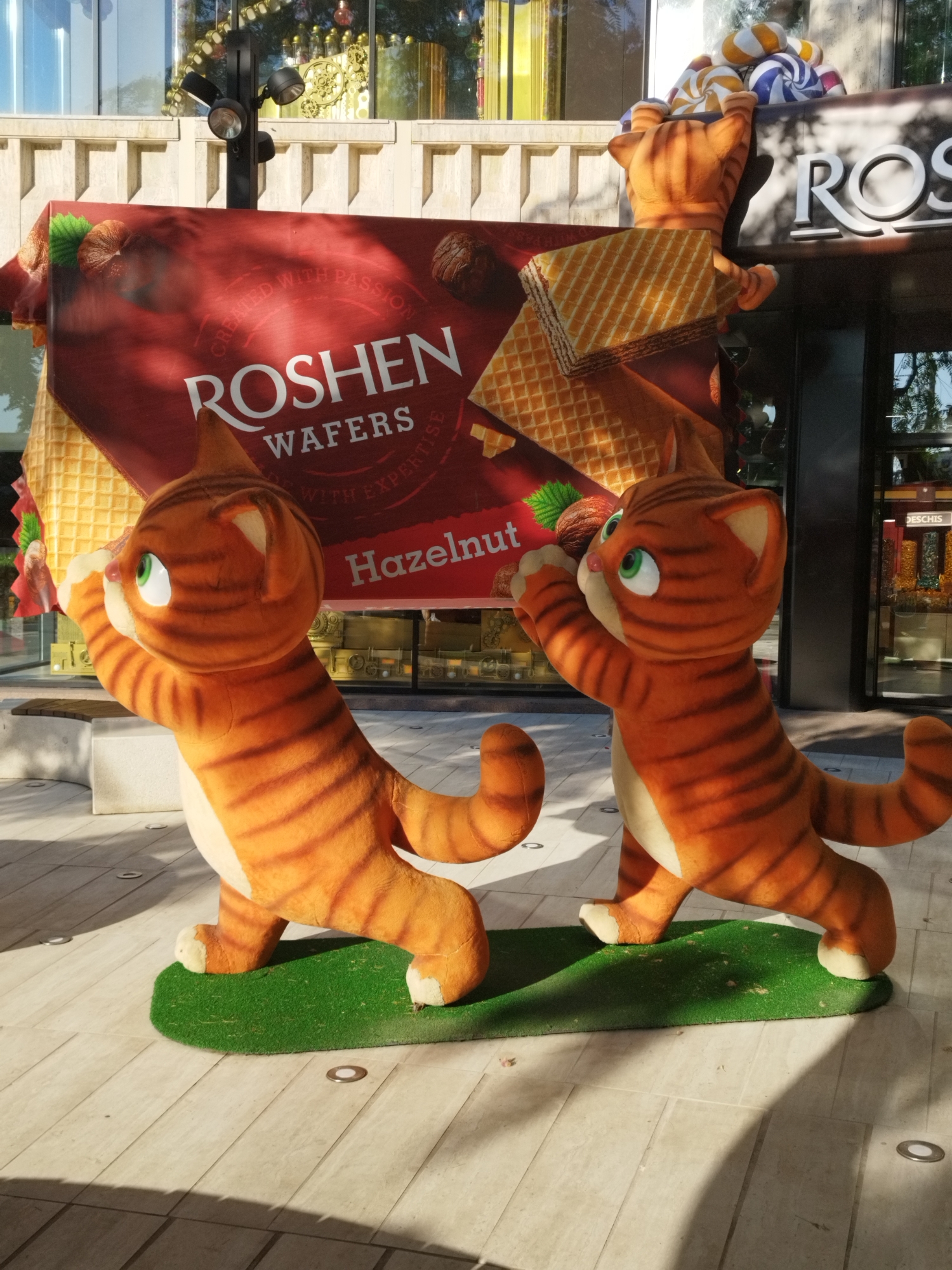 Two plush orange tabby cat figures, each about waist-high to an adult, stand on a green, oval-shaped patch of artificial turf. They are positioned on a light-colored tiled outdoor walkway. The cat on the left faces to the left, while the cat on the right faces to the right. Both cats have wide green eyes, pink noses, and striped bodies with lighter undersides.
Behind the cats is a large red banner advertising "ROSHEN WAFERS WITH EXPERTISE HAZELNUT." The banner features images of hazelnuts, wafers, and the Roshen logo. The background includes a building with large windows and a display of colorful candy. A black pole with three spotlight fixtures is mounted on the building facade. The scene is illuminated by sunlight, casting shadows on the walkway. The location is identified as Bucharest, Romania.
