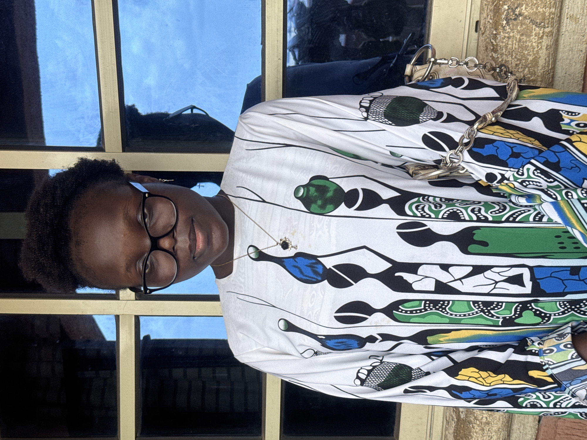 A person wearing eyeglasses and a colorful patterned top stands or sits in front of a window with multiple panes. The window's reflection shows a blue sky. The person also wears a necklace and has a handbag with a silver chain. The environment appears to be outdoors, with part of a building facade visible. The location is inferred to be Onitsha, Nigeria. No specific actions or interactions are apparent in the image.