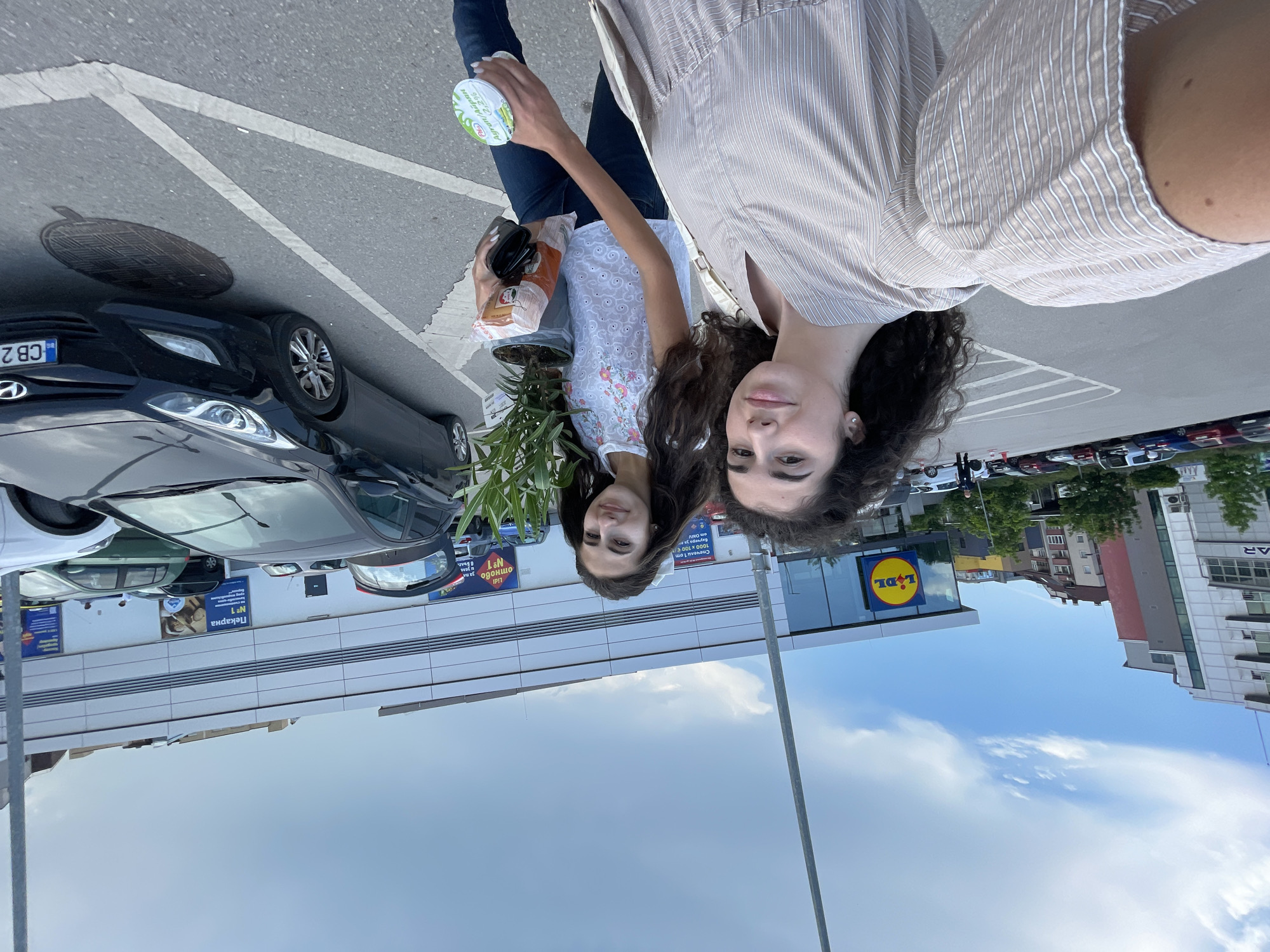 In Sofia, Bulgaria, two young women are pictured on a cloudy day in a parking lot outside a Lidl supermarket. The woman in the foreground, on the left, is wearing a light-brown striped dress and looking at the camera. The woman next to her, on the right, is wearing a white top with floral embroidery and dark jeans. She is holding a potted plant and a plastic bag of groceries. Several cars are parked in the lot, and the Lidl store's signage is visible in the background.