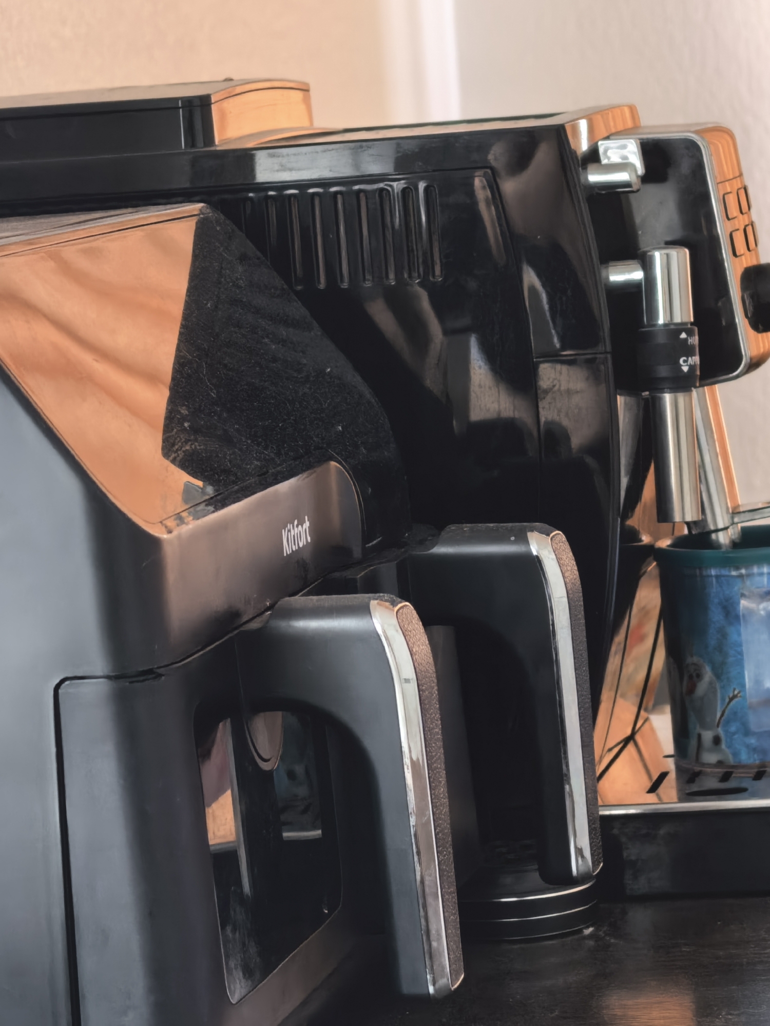The media file displays two kitchen appliances positioned side-by-side on a dark surface, likely a countertop, against a light-colored background wall. The image is taken from a low angle, looking upwards at the appliances, highlighting their glossy surfaces and metallic accents.
On the left, a black appliance, possibly an air fryer, occupies the foreground. It features a sleek, angular design with prominent handle-like elements that have metallic silver edges and textured brown inserts. The brand name "Kitfort" is visible in white lettering on the side of this appliance. Its glossy surface reflects warm, orange-brown tones from the surroundings. Some dust or smudges are apparent on its black finish.
To the right, a black coffee machine is partially visible. It has a metallic steaming wand with a control dial that shows text including "HO" and "CAP". Below the wand, a blue and white mug with a cartoon character, possibly Olaf from Frozen, is placed on a drip tray. The side of the coffee machine also features repeated "CO" text. Similar to the left appliance, its surface is glossy and reflects light, revealing some dust.
No activity is taking place; the appliances appear idle. The lighting suggests an indoor setting, possibly during the day. The overall impression is a snapshot of common kitchen items, showing wear and reflections on their dark surfaces.