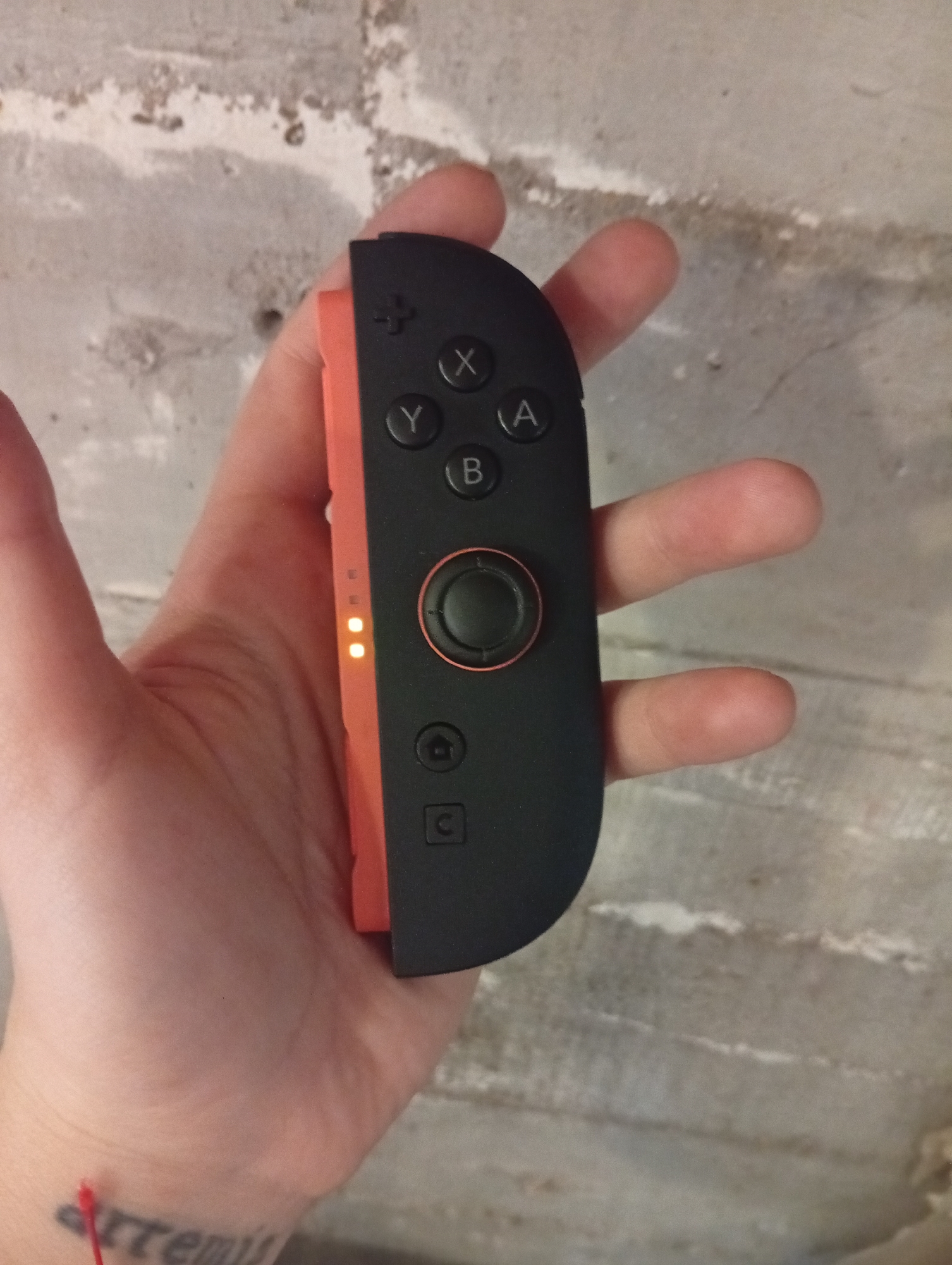 A person's hand is holding a Nintendo Switch Joy-Con controller, specifically the left Joy-Con, against a mottled gray wall. The controller is primarily black with an orange side. Two orange indicator lights are illuminated on the side of the controller. The person's skin is pale. A partial tattoo of the word "artemis" is visible on the wrist, along with what appears to be a red string. The setting appears to be indoors. Location cannot be confirmed to be Buenos Aires, Argentina.