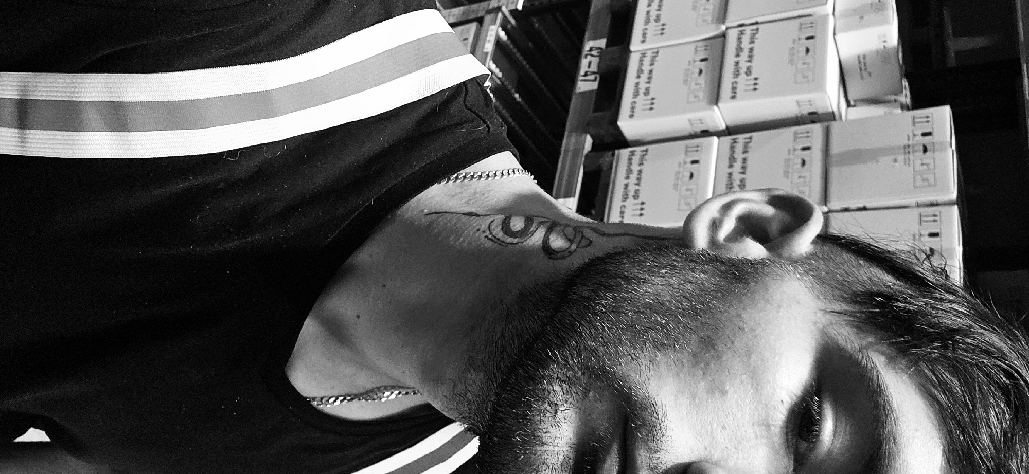 A black and white close-up shot shows the side of a man's face and neck with a tattoo near the ear. He has a chain necklace. The man is wearing a black shirt with white and grey stripes. In the background, boxes are stacked on shelves. Some boxes are labelled "This way up !!! Handle with care" and "42-47." The specific location in Plovdiv, Bulgaria, is indeterminable from the given information.