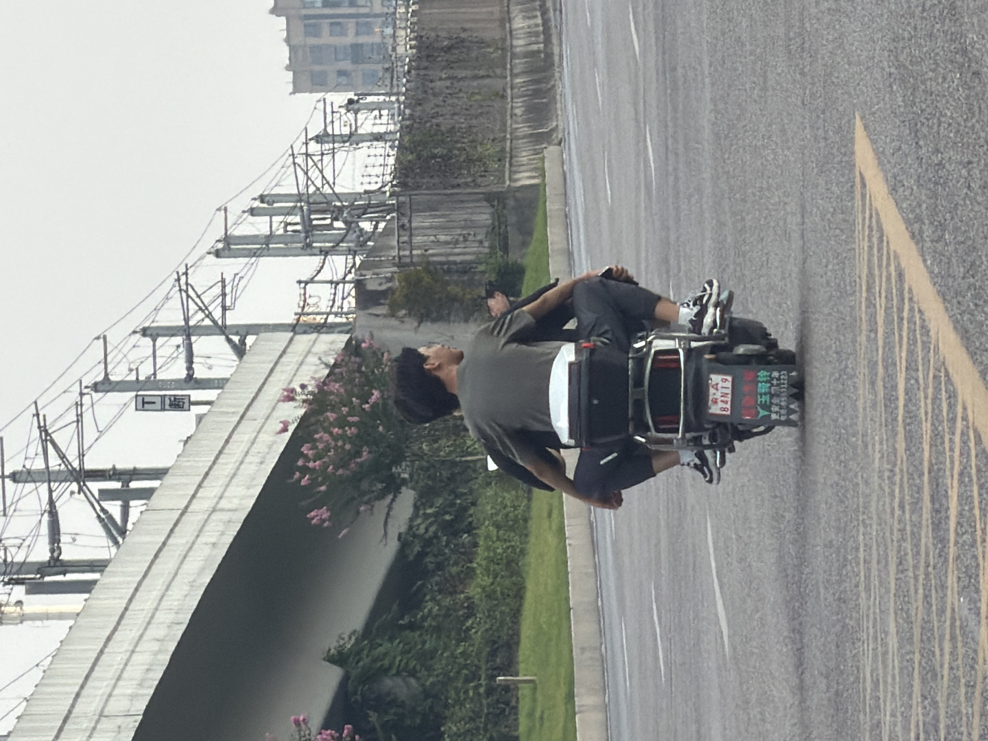 In this image, two individuals are riding on a scooter in Jiangjin District, Chongqing, China. The person in the back is wearing a dark green t-shirt and dark pants, while the person in the front is wearing a black t-shirt and dark pants. They are riding on a paved road with yellow lane markings. In the background, there is an elevated structure that appears to be an overpass or bridge, with overhead power lines and poles. Green vegetation and some pink flowers are visible along the side of the road and under the overpass. A multi-story building is visible in the far background. The license plate on the scooter reads "B4N19" and also contains Chinese characters. The overall scene depicts urban transportation on a cloudy day.