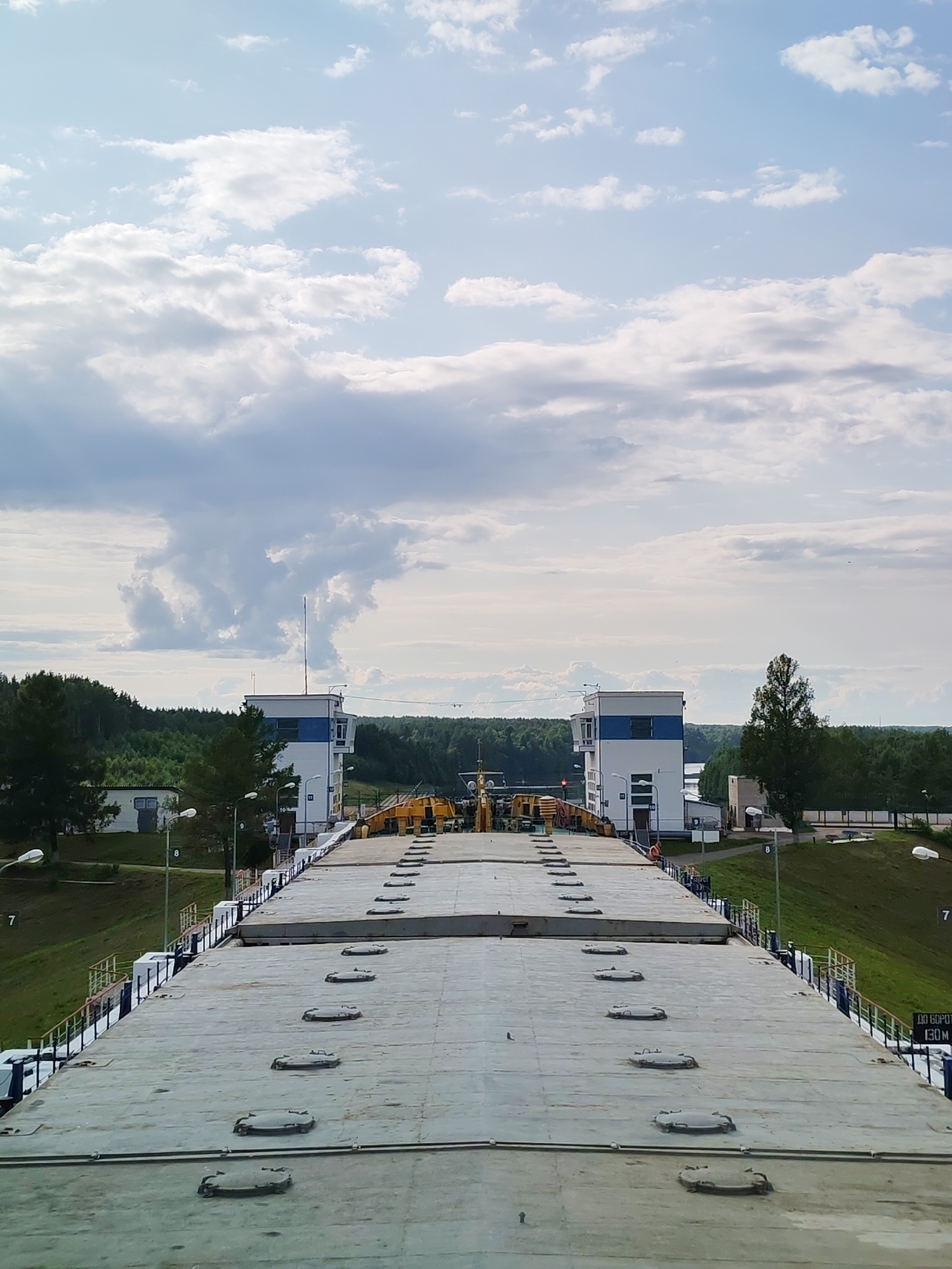 The image depicts a view from the deck of a large vessel, likely a barge or ship, as it approaches a lock system on the Volga-Don Canal in Devyatiny, Russia. The vessel's flat, grey deck fills the foreground, marked by a series of circular hatches. Beyond the vessel, a concrete lock structure with white and blue control buildings is visible. The lock gates appear to be open, allowing passage. Yellow machinery is present on the lock's approach.
The background features a dense, green forest that lines the canal banks. The sky is filled with a mix of light and dark grey clouds, suggesting a partly cloudy day. The overall scene is industrial and functional, characteristic of a major waterway transportation system. There are no people visible in the image. The image is taken from a slightly elevated perspective, looking forward along the canal. A signpost with Cyrillic text, "ДО БОРОТА 130М" (To the Lock 130m), is visible on the right side of the frame.