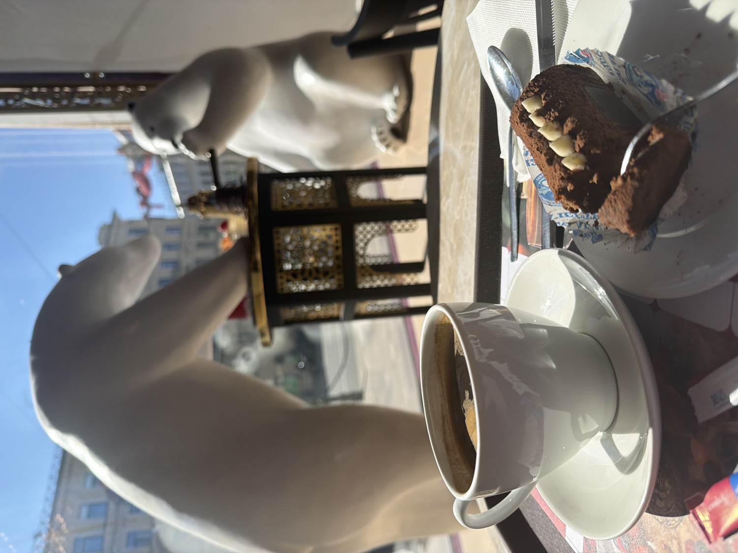 This media file captures a bright daytime scene inside what appears to be a cafe or restaurant, offering a view of an urban landscape through a large window.
In the foreground, a white ceramic coffee cup filled with dark coffee sits on a matching saucer on a patterned placemat. Beside it, a slice of dark chocolate cake, possibly a brownie or similar dessert, rests on a white plate with a fork already inserted into it. The cake features distinctive white rectangular details on its top surface, resembling teeth or pieces of white chocolate. The cake is partially wrapped in paper with blue patterns and the visible text "ALEXANDRIA". Further back on the table, a spoon and a napkin are visible.
Through the large window, two white polar bear figurines or statues are prominent. The closest polar bear stands with its front paws seemingly supporting a decorative, ornate lantern with dark frames and gold-colored filigree patterns. A second, more distant polar bear figurine is also visible. Beyond the window and the figurines, a clear blue sky is visible above urban buildings, suggesting good weather. The buildings in the background are somewhat blurred, indicating the scene is focused on the interior and the window display.
The strong, natural light streaming through the window suggests it is daytime, likely morning or early afternoon, casting distinct shadows on the table and objects. The overall atmosphere appears calm and relaxed, characteristic of a cafe visit. The presence of the polar bear figurines and the general aesthetic hint at a unique establishment within Saint Petersburg, Russia.