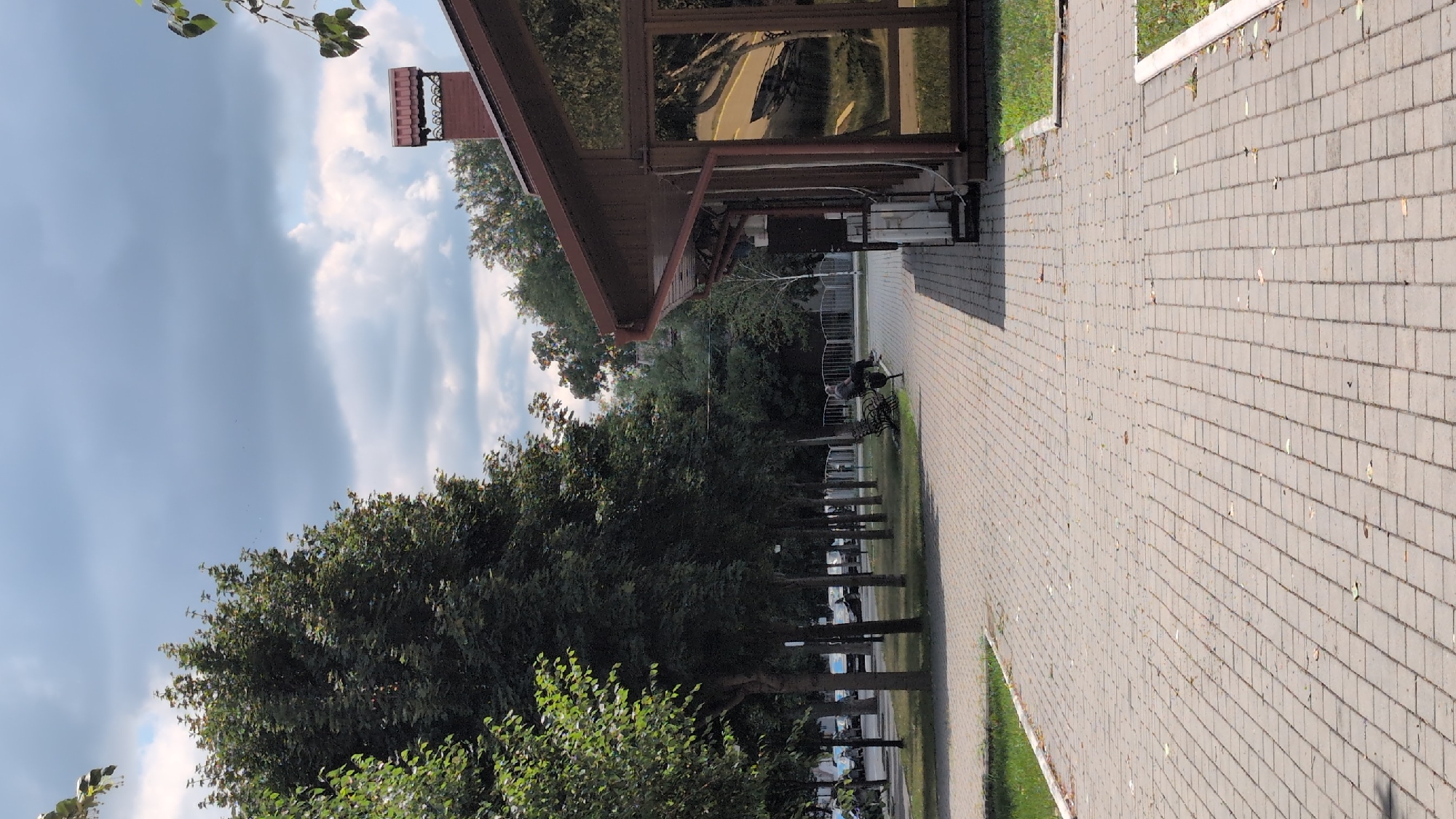 A low-angle, vertical full shot captures a paved walkway with a person seated on a bench to the left, observing a brown building with large windows and a brick chimney to the right. The scene is set in Izmail, Ukraine, under a cloudy sky.
The walkway, made of gray pavers, stretches from the foreground into the distance, bordered by a grassy area on the left and the building on the right. Trees with lush green foliage line the left side of the walkway, casting shadows on the path. Beyond the trees, parked cars are visible.
A solitary figure, dressed in dark clothing, is seated on a dark-colored bench, facing towards the building. The figure's posture suggests a moment of rest or observation.
The building, constructed of wood, features a brown roof and large, reflective windows that mirror the surrounding trees and sky. A red brick chimney with decorative elements rises from the roof. The building's exterior appears well-maintained.
The sky is filled with a mix of gray clouds and patches of blue, indicating overcast conditions. The overall atmosphere is serene and quiet, suggesting a peaceful day in this urban setting.