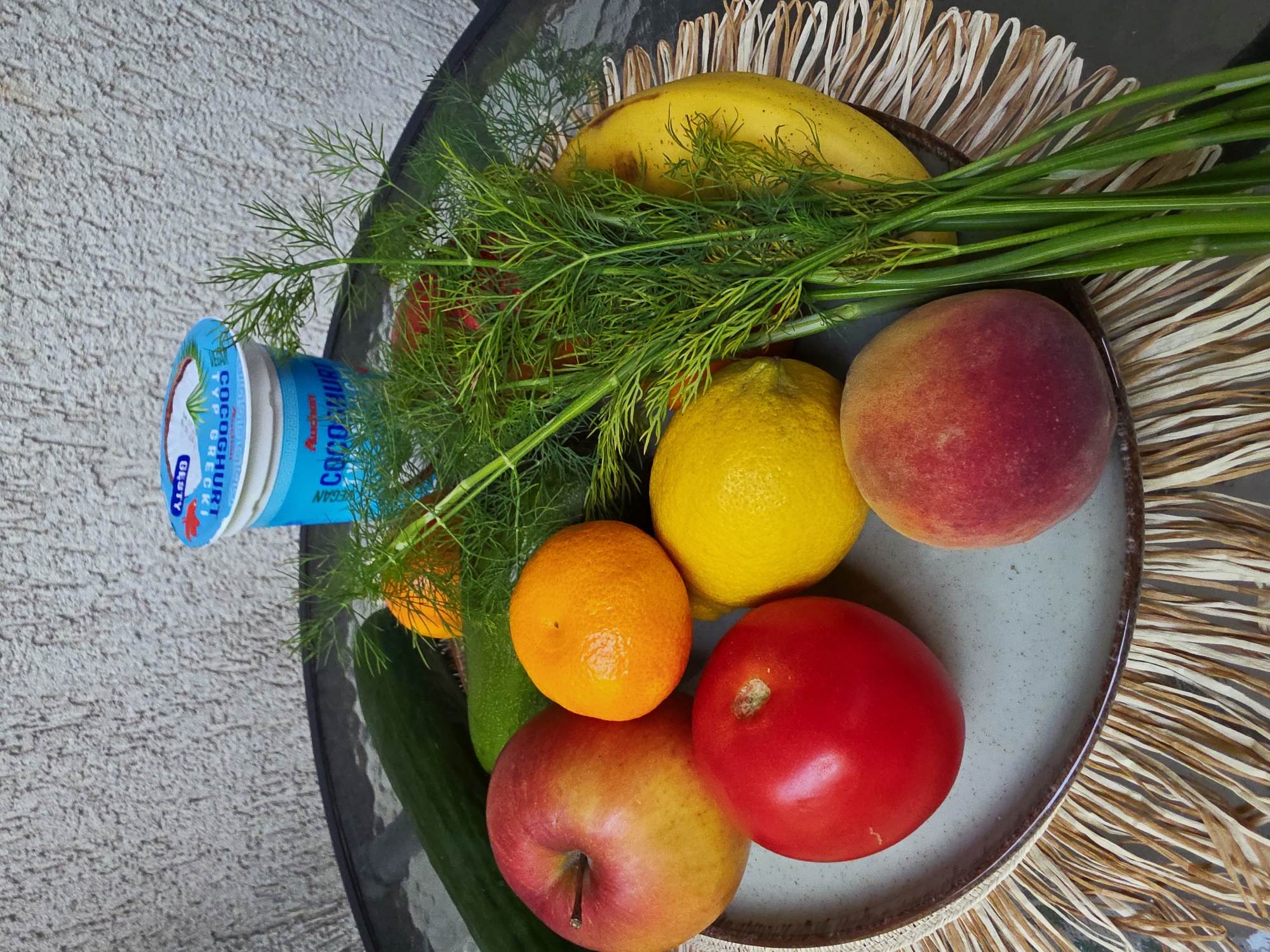 A top-down, slightly angled shot captures a vibrant assortment of fresh produce and a food container arranged on and around a light gray, speckled plate. The plate sits on a round, dark glass table, beneath which a light brown, woven placemat with fringed edges is visible.
Dominating the center of the image is a fresh bunch of green dill, splayed across the plate and extending towards the upper right. On the plate itself, from left to right, are a yellow lemon, a bright orange, a red and yellow apple, and a solid red tomato. To the right of the plate, partially on it, are a second yellow lemon and a reddish-yellow peach. Another peach, similar in color, is located just above the second lemon. A yellow banana is visible behind the dill, near the top edge of the plate.
To the left of the plate, a long green cucumber rests, with a portion of a blue and white container positioned vertically beside it. The container has text in both Cyrillic and English, clearly showing "Auchan" as the brand and "COCONUT" and "VEGAN" indicating its contents. Other visible text includes "COCOCNUHT", "TYP GR E C", and "GLA".
The background features a textured, light gray wall, suggesting an outdoor setting like a patio or balcony. The scene is well-lit, indicating daylight conditions.
