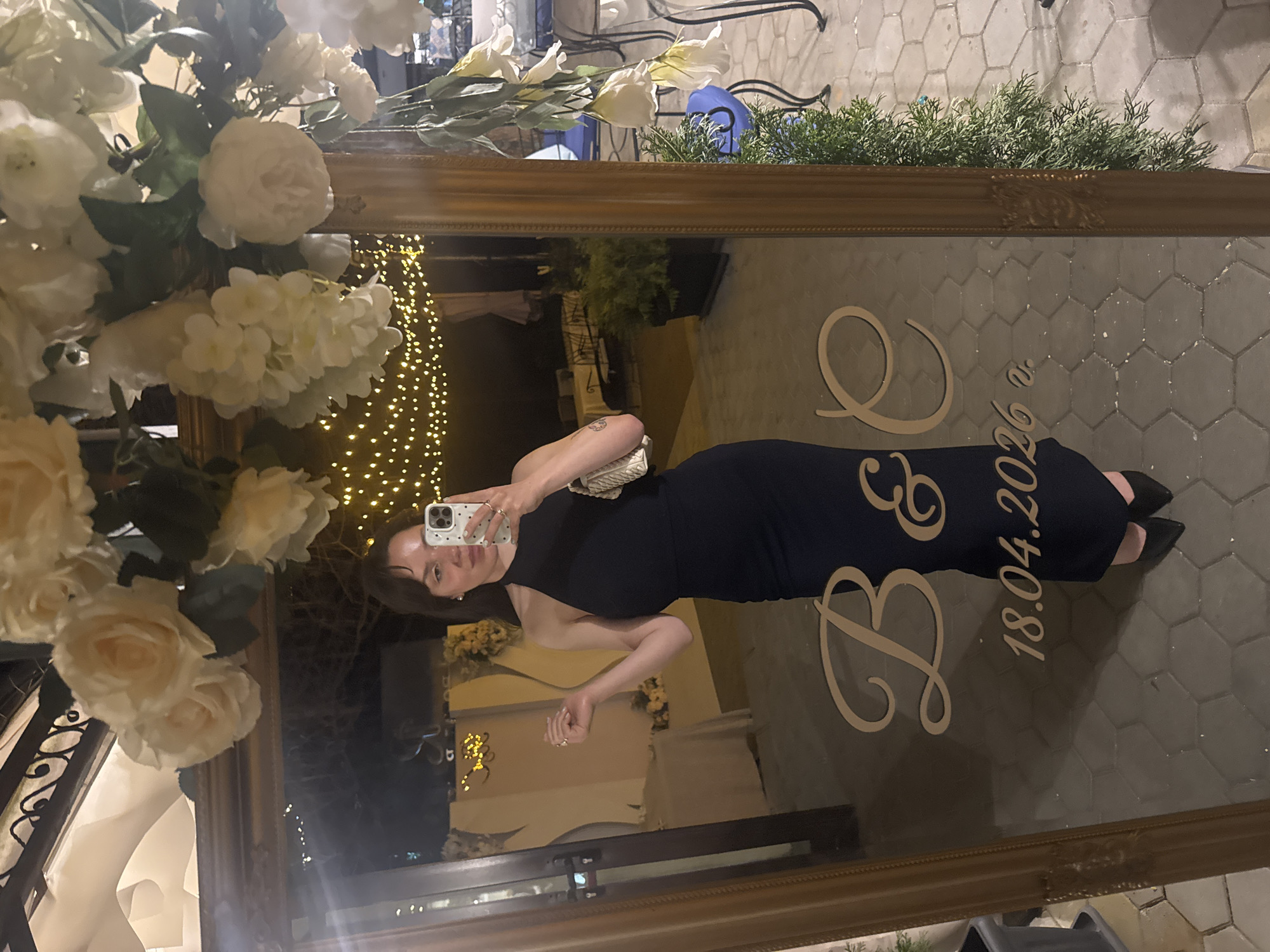 A woman is taking a selfie in a large mirror in Blagoevgrad, Bulgaria. The mirror is decorated with white flowers and greenery. Reflected in the mirror are outdoor lights and buildings. In the reflection, the woman is wearing a dark dress and holding a phone. The mirror also has lettering etched into it that reads "B & E 18.04.2026". The ground surrounding the mirror appears to be paved with hexagonal tiles.