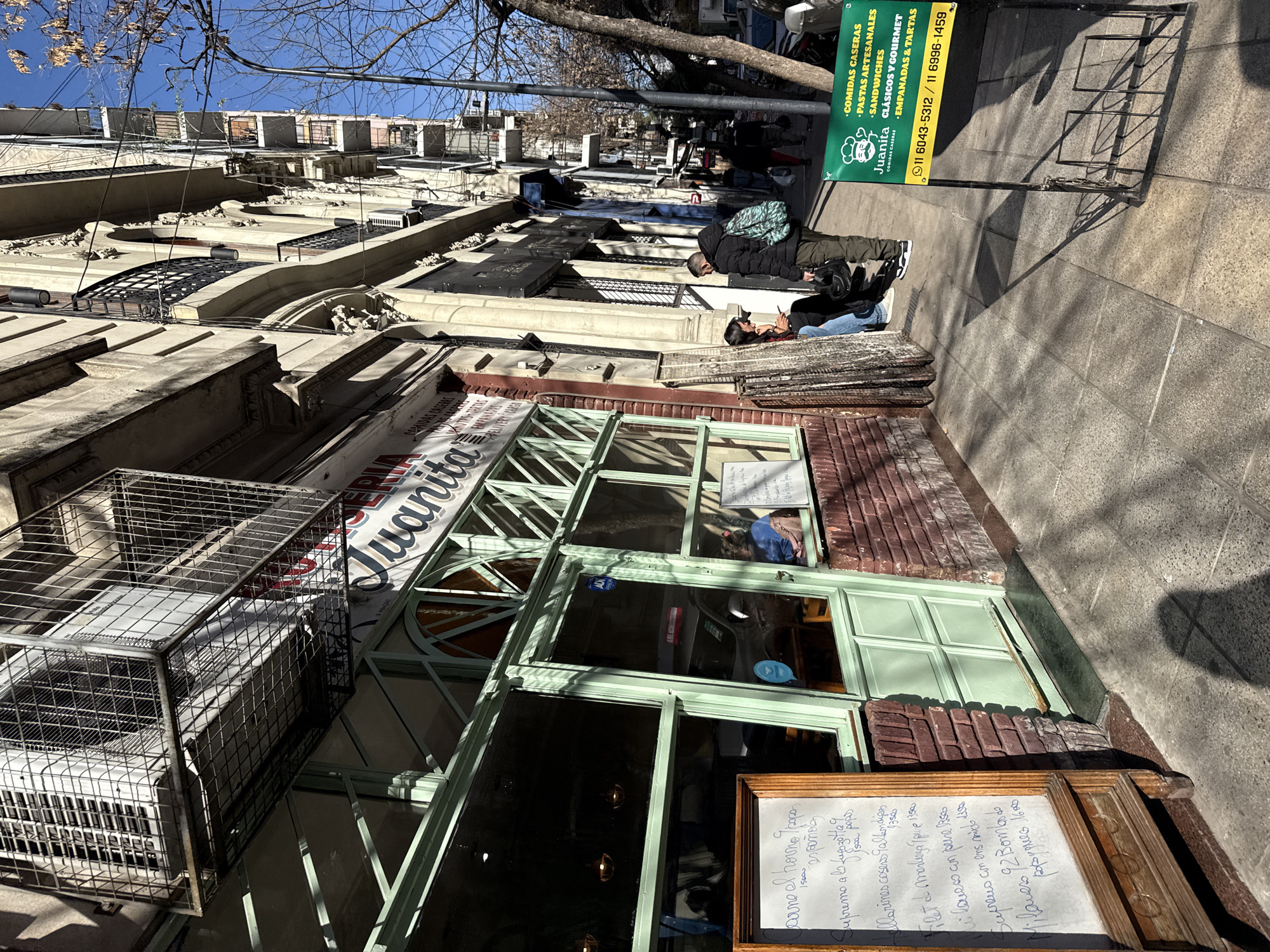This image captures a bright, sunny day on a city street in Buenos Aires, Argentina, featuring a storefront named "Juanita." The scene is bustling with everyday activity under a clear blue sky.
The main subject is the "Juanita" establishment, which appears to be a cafe or restaurant, indicated by its menu and signage. The storefront features large glass windows with light green frames and a brick facade. To the left, an air conditioning unit is housed within a protective metal cage. On the sidewalk, a wooden chalkboard sign displays a menu in handwritten Spanish, listing "COMIDAS CASERAS" (Homemade Food) and various dishes with prices, such as "Supremo a la Napolitana," "Milanesas caseras con Albondigas," "Filet de merluza con puré 1350," and "Milanesa de bondiola de cerdo con papas fritas 1600." A green banner sign, also for "Juanita," hangs near the street, advertising "PASTAS ARTESANALES," "SANDWICHES CLÁSICOS Y GOURMET," and "EMPANADAS & TARTAS," along with two phone numbers: "11 6043-5312 / 11 6996-1459."
Two individuals are visible on the sidewalk in front of the shop. One person is seated on a low ledge, wearing a dark jacket and jeans. Another person stands nearby, dressed in a dark jacket and carrying a green patterned backpack. Both appear to be casually waiting or observing.
The setting reveals an urban environment with multi-story buildings lining the street, showcasing classical architectural elements like balconies and ornate details. Bare tree branches are visible against the blue sky, suggesting late autumn or winter. Strong sunlight casts distinct shadows across the pavement and buildings, indicating mid-morning or afternoon. A metal bicycle rack stands empty on the sidewalk.