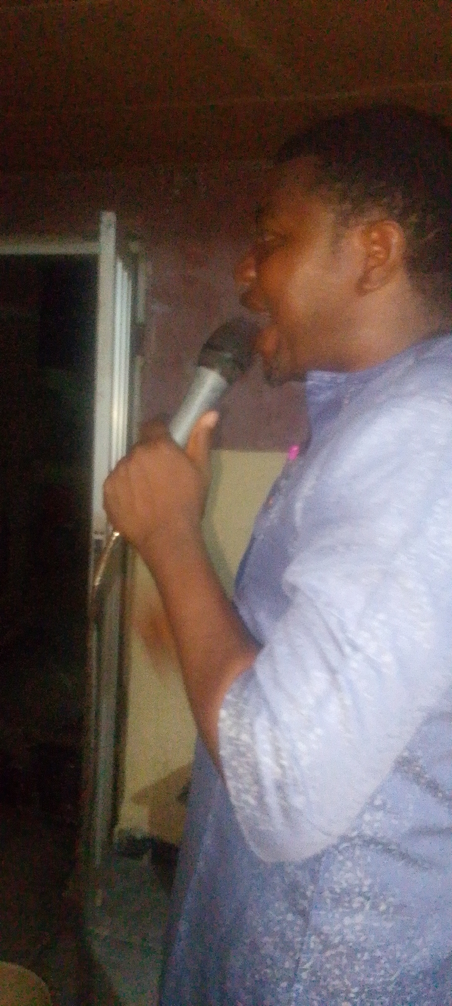 A medium shot shows a man in a light blue patterned shirt holding a microphone to his mouth. The man is facing to the left with his mouth open as if singing or speaking. He is dark-skinned with short, dark hair. The background is dark and out of focus, showing a doorway on the left. Given that the location is Lokoja, Nigeria, the environment is likely indoors, perhaps a small event or gathering space.