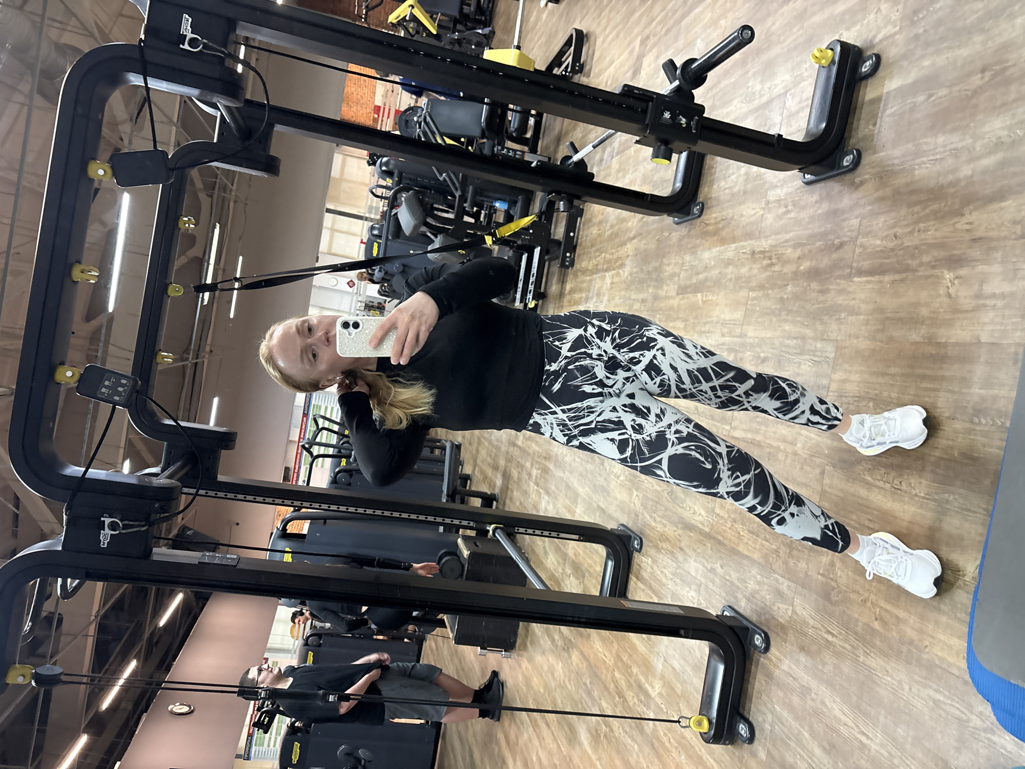The image shows a gym interior. A woman in black and white leggings and a black shirt is taking a selfie in a mirrored exercise machine. The mirror reflects the gym environment, revealing other gym equipment and a man exercising. The gym has a wooden floor. The location is Chaikovsky, Russia.