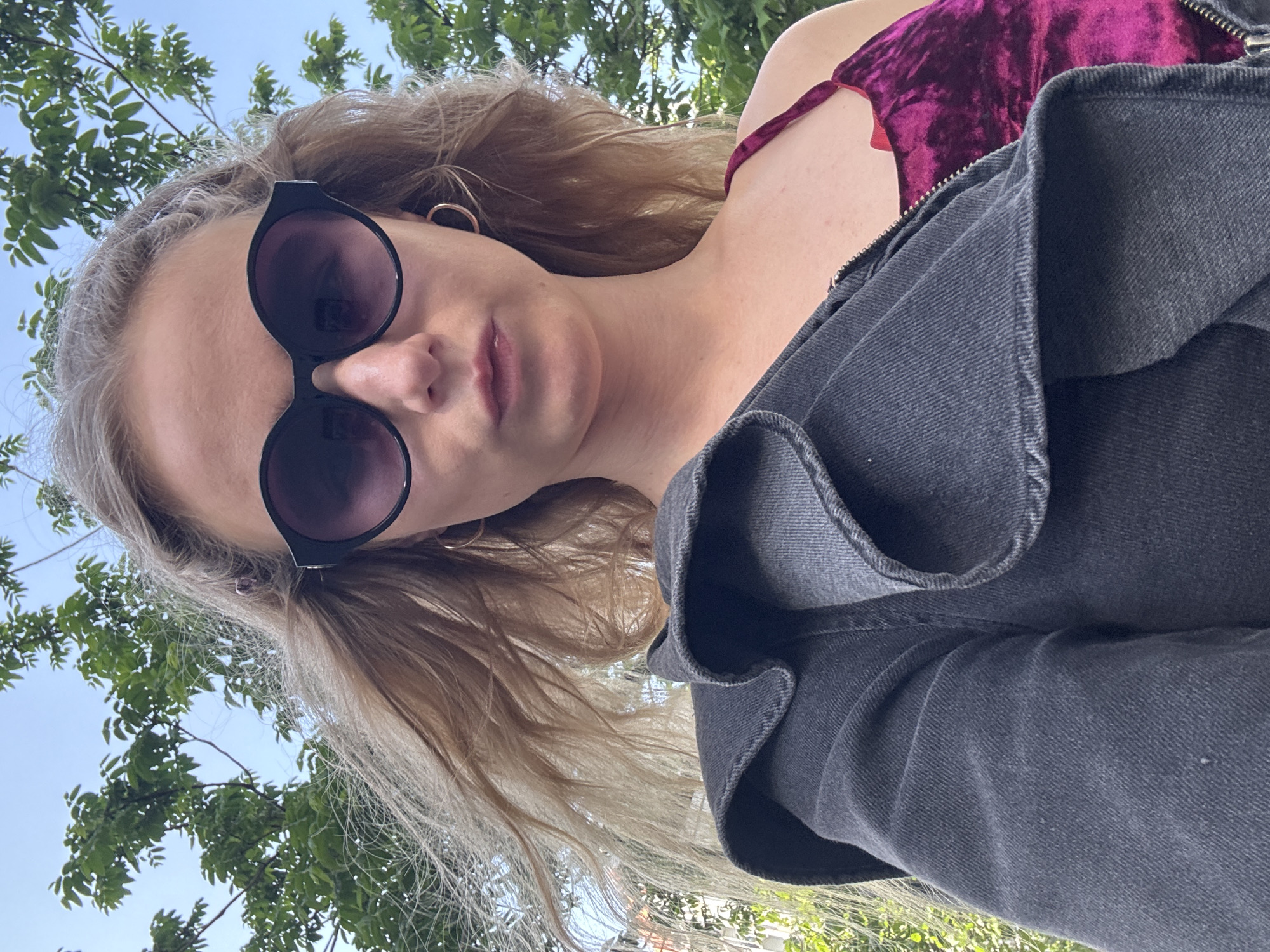 A low-angle shot of a woman with light blonde hair wearing round, dark sunglasses. She has on a dark gray jacket over a purple velvet top. The background shows green leafy trees and a light blue sky. The location is in a city, as a building can be seen through the trees.