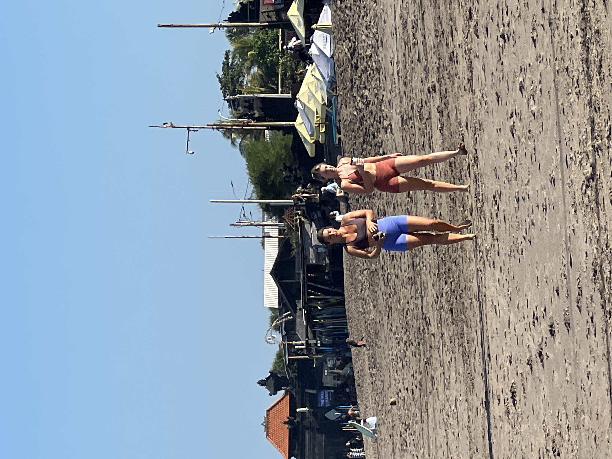 Two young women walk on a sandy beach on a sunny day. The woman on the left wears a dark sports bra and blue shorts, and the woman on the right wears a rust-colored sports bra and shorts. They are walking towards the right side of the frame. In the background, there are several buildings with terracotta roofs, market stalls, beach umbrellas, and surfboards. The sky is clear and blue. The scene appears to be in North Kuta, Indonesia.