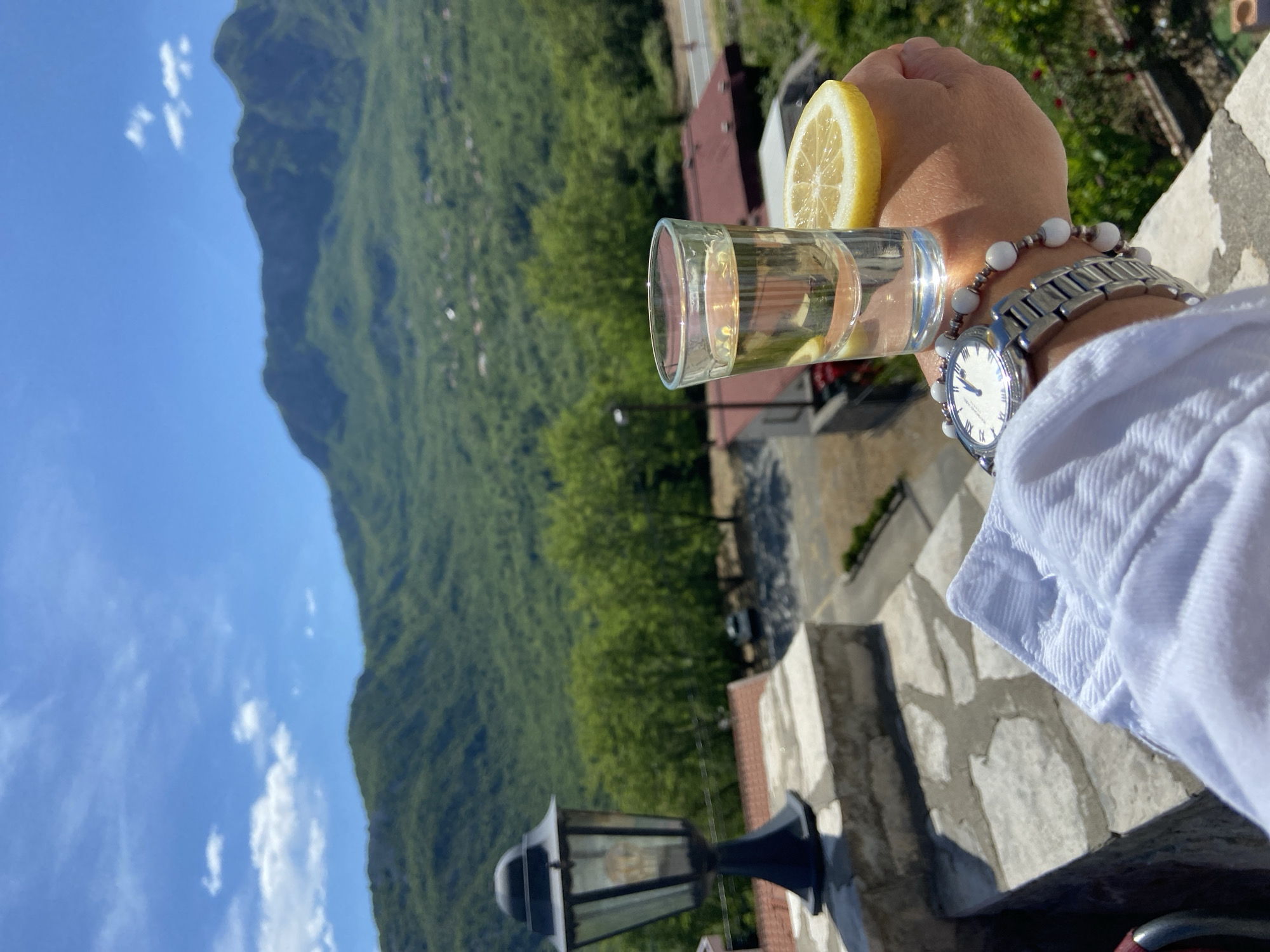 A person is holding a shot glass with a lemon slice attached, positioned in front of a mountainous landscape in Sotonići, Montenegro. The person is wearing a white shirt, a silver watch, and beaded bracelets. The background features a forested mountain under a blue sky with scattered clouds. A lamp post and parts of stone structures are visible, suggesting a terrace or elevated area.