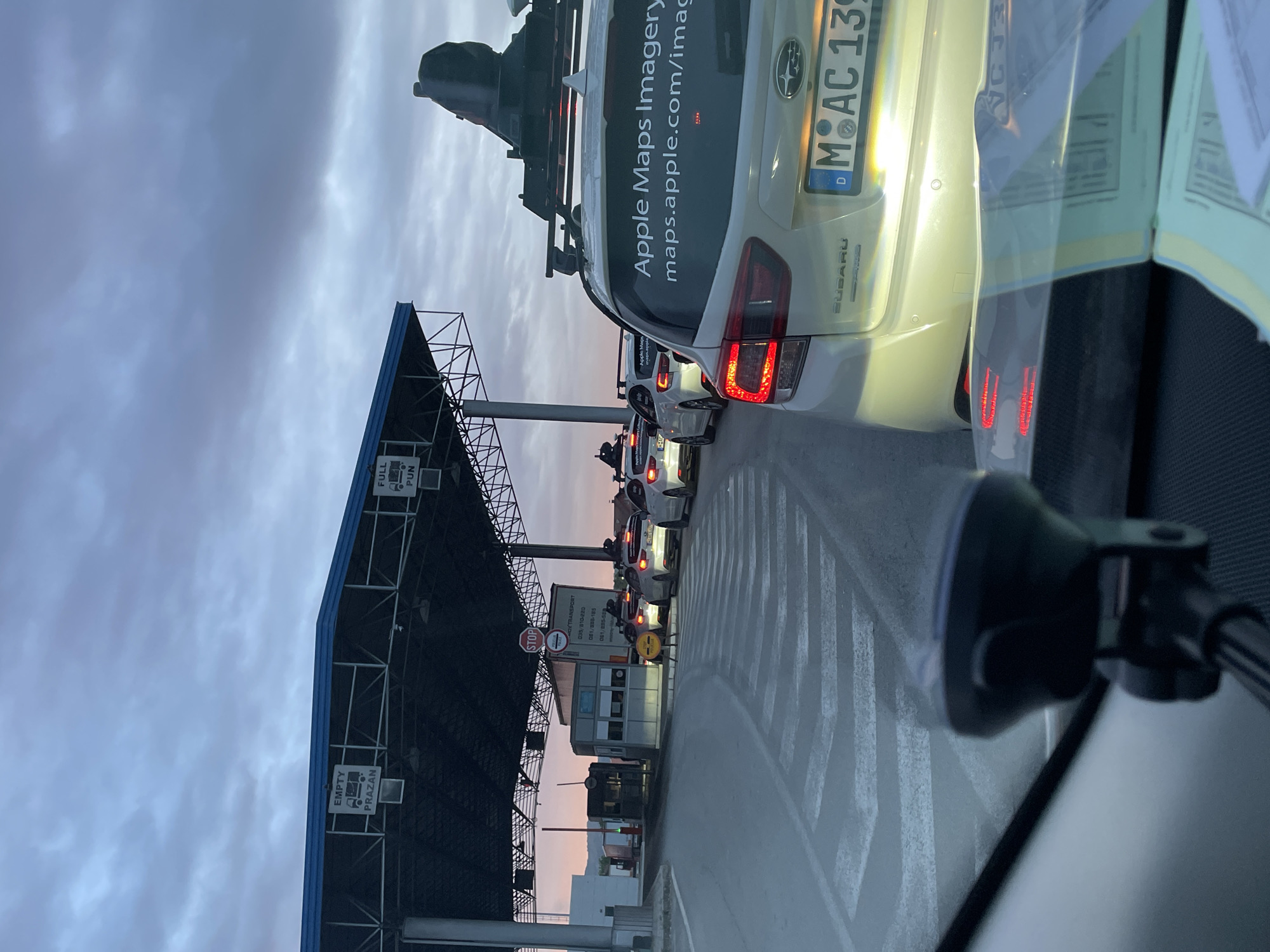 The image depicts a line of vehicles, including one with "Apple Maps Imagery" signage, at a border crossing. The crossing structure has signs reading "EMPTY/PRAZAN" and "FULL/PUN," suggesting a customs or toll point. A "STOP" sign is visible. The car in front has a German license plate, "M-AC 139," indicating it is registered in Munich. The scene appears to be in Karakaj, Bosnia and Herzegovina based on the "PRAZAN" sign and possible truck transport vehicle. The sky is cloudy.