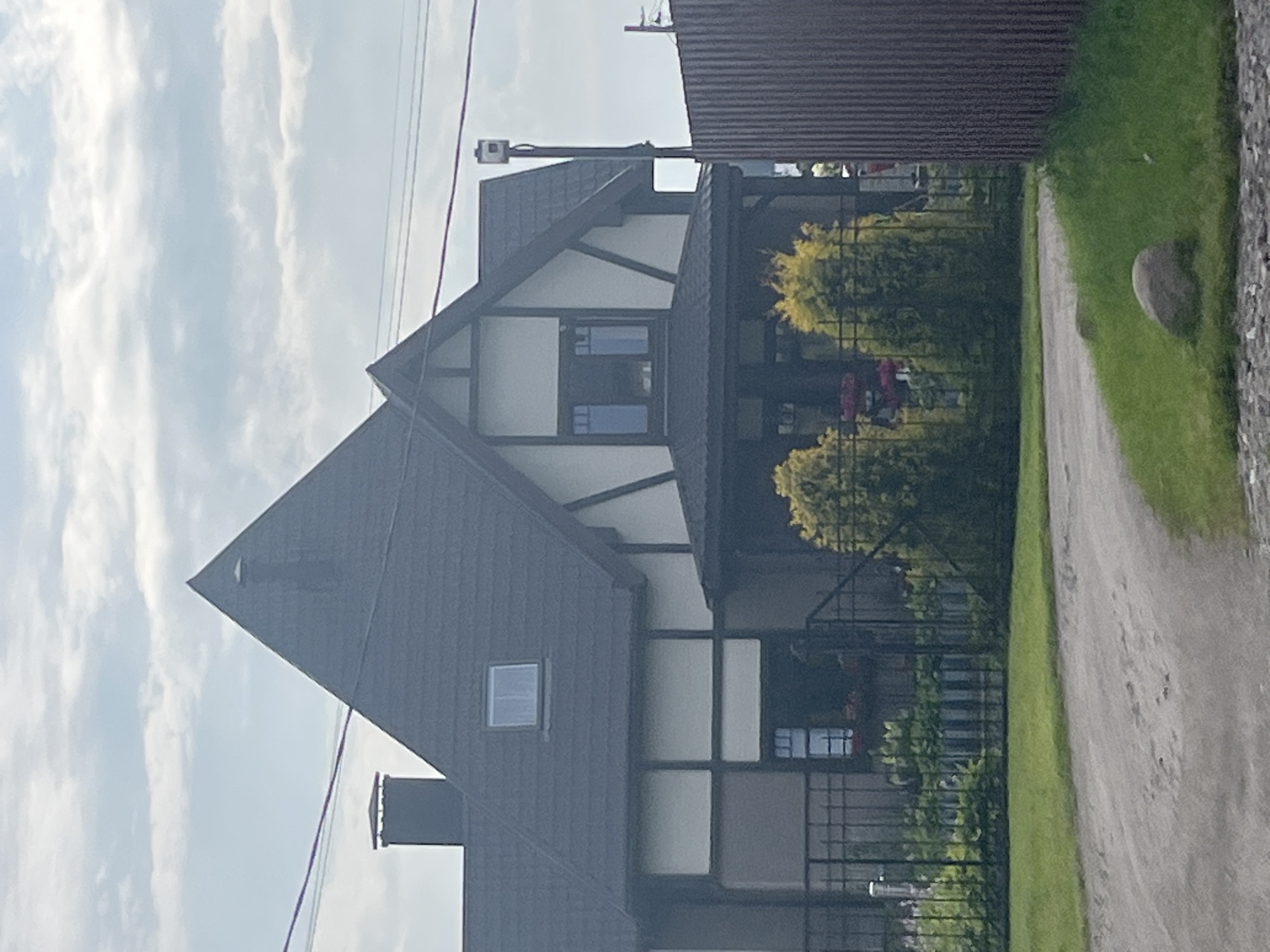 The image shows a two-story house with a dark grey, steeply pitched roof in the city of Babigonskaya Valley, Russia. The house has a light-colored facade with dark trim and timber-style detailing. There are several windows visible, including a small square window on the upper roofline and a larger rectangular window on the upper floor. A dark-colored chimney protrudes from the left side of the roof. A covered porch area with dark columns is situated at the front entrance. In front of the house, a black metal fence encloses a garden with lush green plants and some flowering shrubs. Two conical evergreen trees are positioned in front of the fence. To the right of the house, a tall, dark fence made of corrugated metal runs along the property line. The foreground features a dirt driveway curving to the left, with a grassy area on the right containing a large grey rock. The sky above is partly cloudy. A security camera is mounted on a pole to the right of the house.