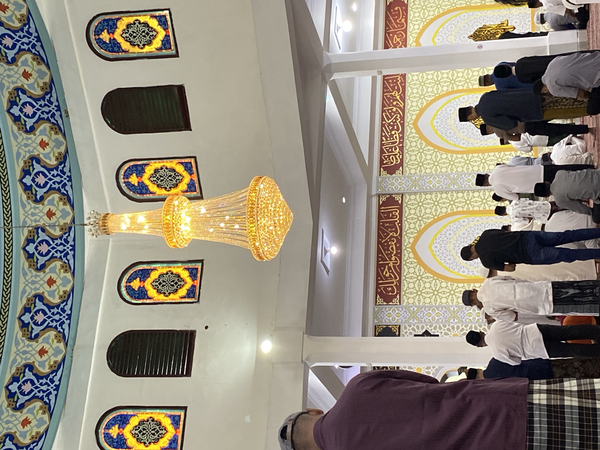 The image depicts the interior of the Grand Mosque of Lhokseumawe, Indonesia, during prayer. A group of men, dressed in traditional attire such as sarongs and caps, are engaged in prayer. The mosque's interior features ornate decorations, including intricate patterns on the walls and stained-glass windows with Islamic motifs. A large, elaborate chandelier hangs from the ceiling, illuminating the space. The architecture suggests a significant religious building, characteristic of mosques in the region. The overall atmosphere is one of solemnity and devotion.