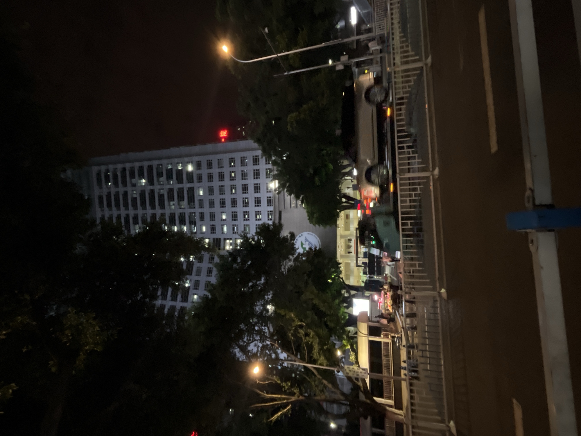A nighttime exterior scene in Yuzhong District, China, shows a tall, illuminated multi-story building with rows of lit windows. In the foreground, a street is visible with a white metal barrier. A light-colored SUV is driving on the road, and a green vehicle, possibly a truck or van, is parked nearby. Several trees with dark foliage are present, and a street lamp with a visible light bulb illuminates the scene. A large circular logo is visible on the building's facade. Red Chinese characters are also present on the building. The sky is dark.