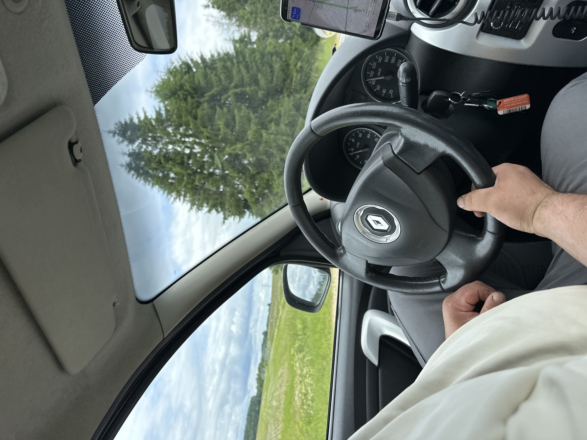 A man's hands are on the steering wheel of a Renault car as he drives through a rural area. The car's interior is visible, including the dashboard with a GPS navigation system on a phone mount and a set of keys hanging from the ignition. Outside the car, a green field with scattered white flowers stretches towards a line of trees under a cloudy sky. The scene suggests a drive through the countryside, possibly in the vicinity of Ilyanskiy selskiy Sovet, Belarus, given the rural landscape.