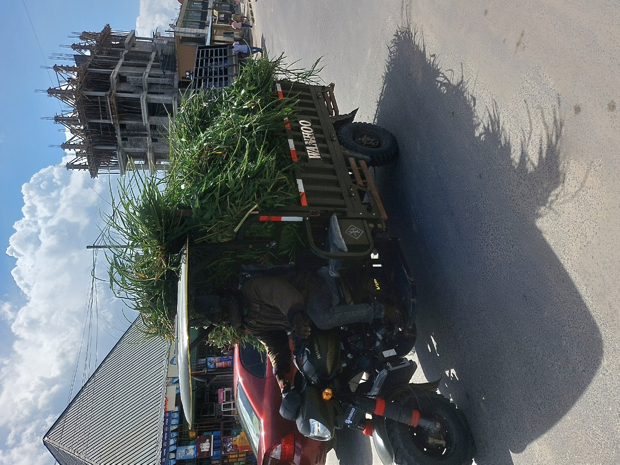 This image captures a vibrant street scene in an urban environment, likely in Dar es-Salaam, Tanzania, during the bright hours of late morning or early afternoon, as evidenced by the distinct shadows cast on the road.
The primary subject is a person riding a dark-colored motor-tricycle, which is heavily loaded with a substantial pile of tall, green, leafy plant material, possibly fodder or produce. The cargo bed of the tricycle clearly displays the brand name "WANHOO" in white lettering, with reflective red and white strips. The "WANHOO" logo is also visible on the motorcycle's fuel tank, and "VIP" can be seen towards the rear of the vehicle.
In the background to the right, a multi-story concrete building under construction is prominent. It features exposed concrete slabs and columns, along with extensive wooden formwork and rebar projecting upwards, indicating active development. To the left, a red car is partially visible, either parked or stopped next to the motor-tricycle. Behind the car, a building with a silver corrugated roof houses what appears to be a shop, with shelves displaying various products, including bottles that seem to bear "Pepsi" logos.
The sky is bright blue with large, white, fluffy cumulus clouds. The road surface is paved. The elongated shadows of the motor-tricycle and its cargo stretch across the asphalt, adding depth and a sense of movement to the scene. The image illustrates a practical mode of transport for bulk goods amidst ongoing urban development and commercial activity.