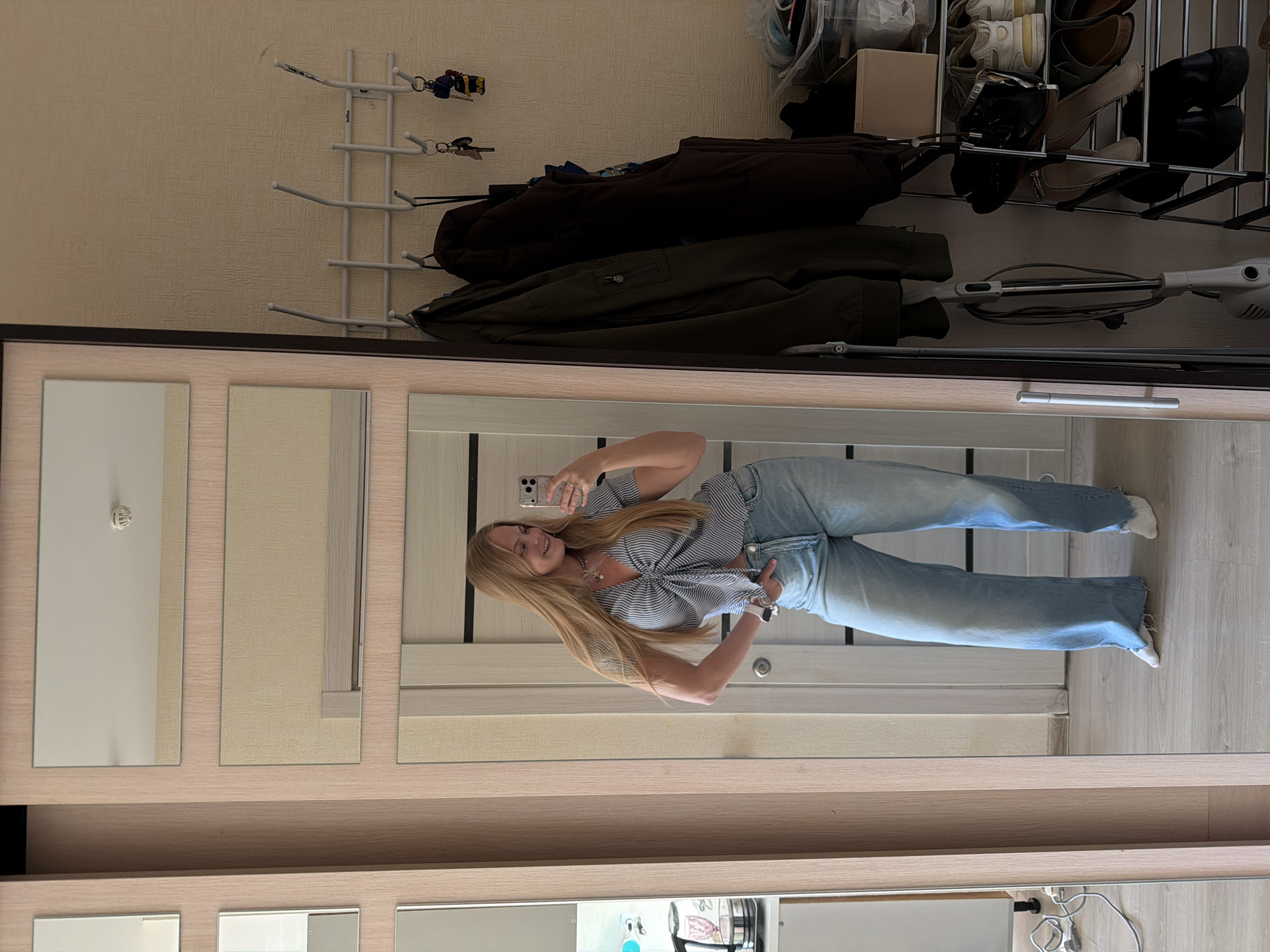 A young woman is captured in a full-length mirror selfie, smiling and holding her smartphone to take the picture. She has long, straight blonde hair and is wearing a blue and white striped crop top with a tie-front detail, light blue loose-fitting jeans, and white socks.
The mirror, part of a light-toned wooden furniture unit possibly a wardrobe or hallway storage, reflects the domestic setting behind her. On the wall in the reflection, a white wall-mounted coat rack holds several sets of keys and dark-colored jackets or coats. The word "BalDog" is faintly visible on the coat rack. Below the coats, a shoe rack displays various pairs of shoes, including heels, flats, and sneakers. A white stick vacuum cleaner is also visible, leaning against the wall beneath the shoe rack. The wall itself is light-colored with a subtle textured pattern, and the floor appears to be light wood or laminate. The setting is an indoor space, likely an entryway or hallway, in Izhevsk, Russia, characterized by neat organization and casual living.
