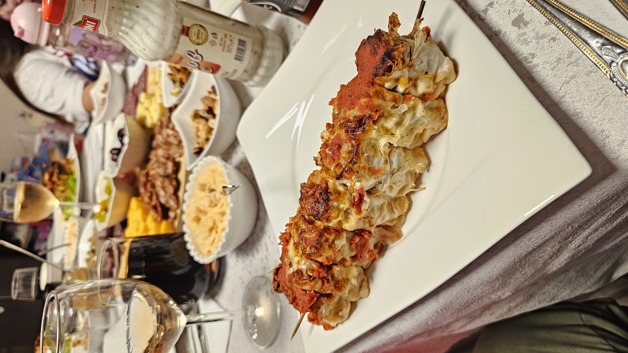 The image depicts a table setting, likely indoors, with various food items and dinnerware. A prominent dish is a kebab of dumplings or pasta with tomato sauce, presented on a square white plate. In the background, a variety of other food items are visible, including what appears to be a charcuterie board with cheese, meats, and spreads. There are also glass bottles and several wine glasses, suggesting a meal with wine. The location is not discernible based solely on the visual information. It is not possible to determine if this is in Varna, Bulgaria, without additional context or identifying features.