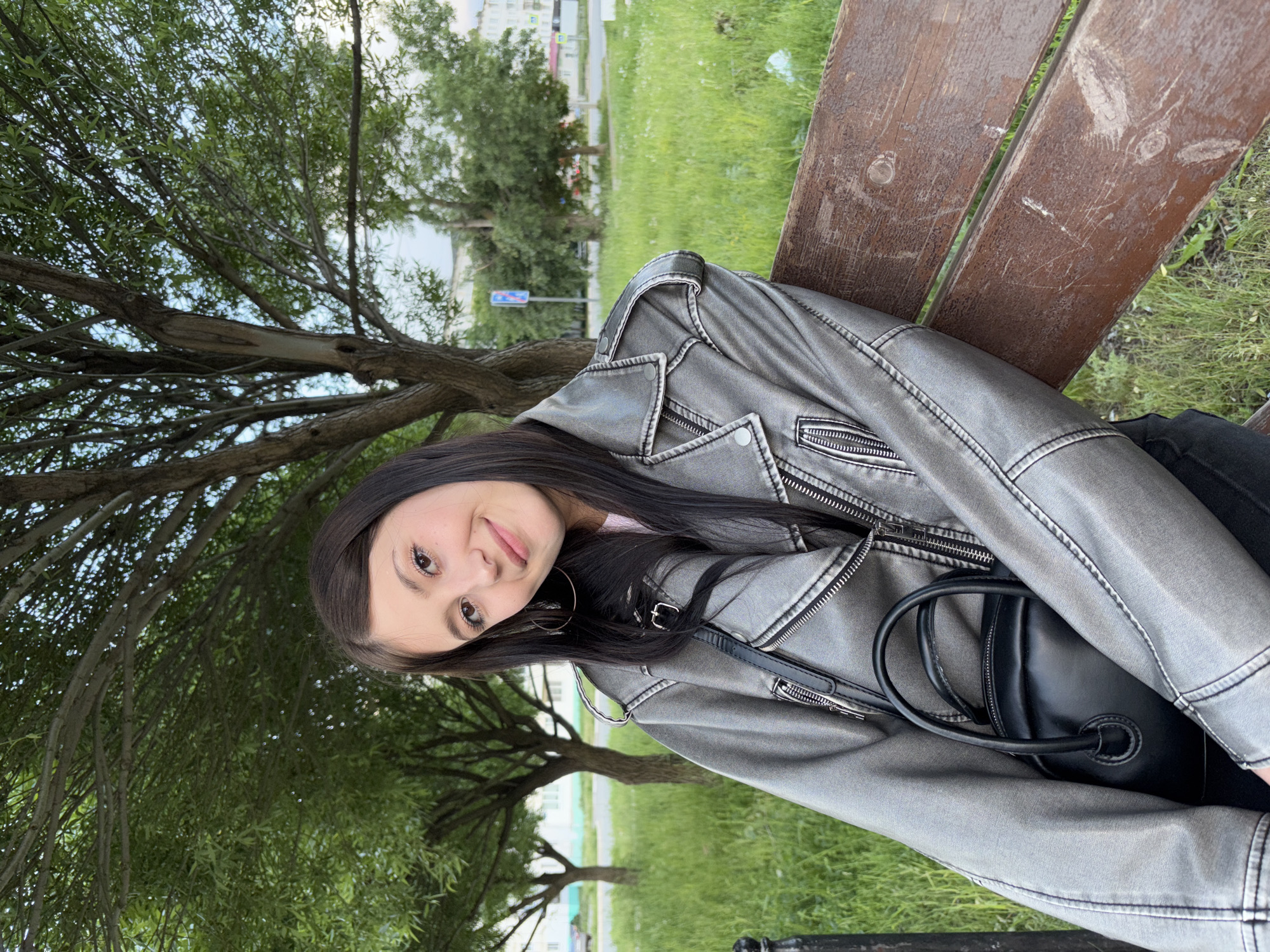 A young woman with dark hair is sitting on a wooden park bench. She is wearing a grey leather jacket and has a black purse. The background includes a tree and a grassy area. In the distance, there is a building, suggesting the location is a city park. The scene is in Chaikovsky, Russia.