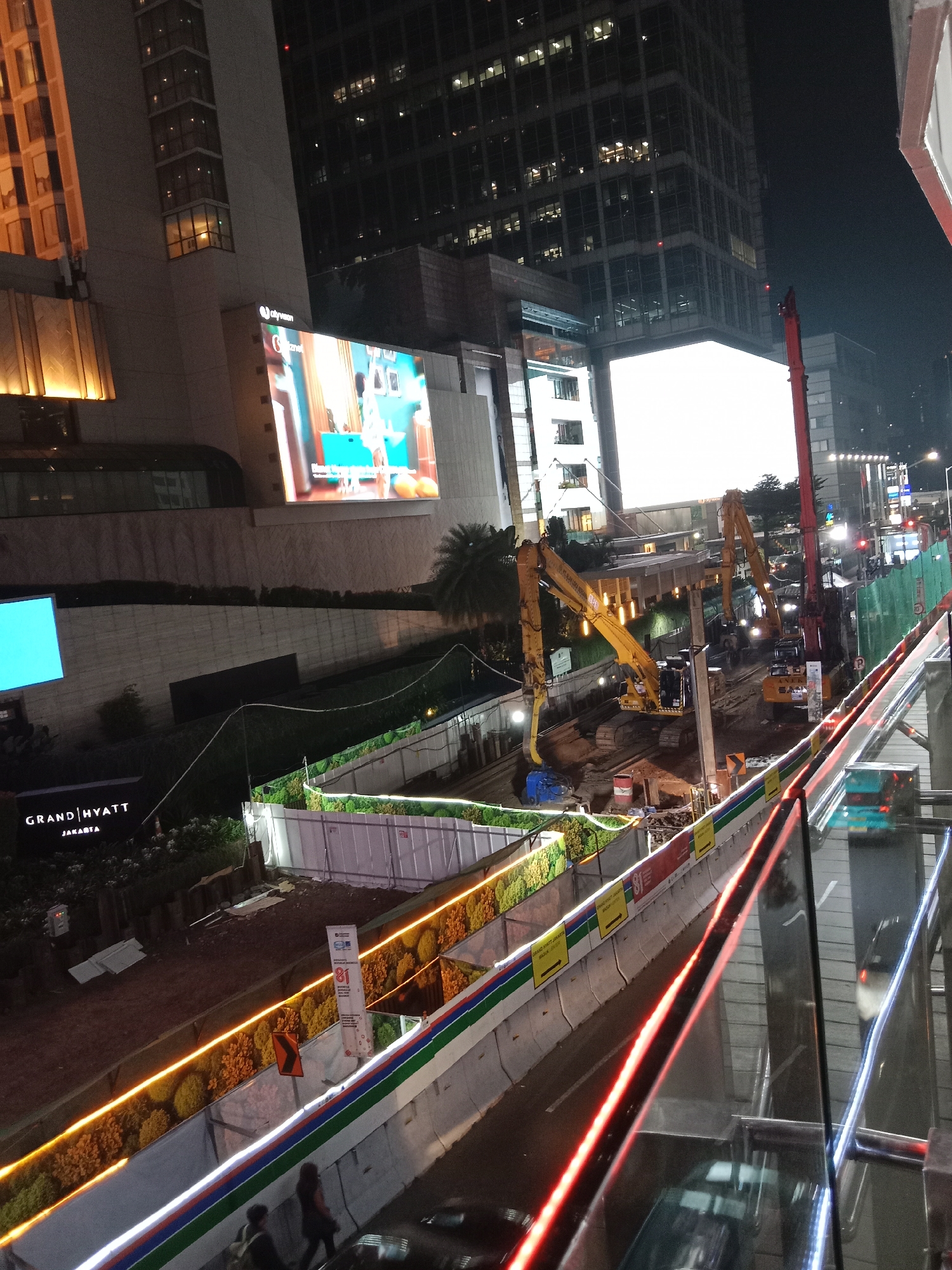 The image shows a construction site at night in Central Jakarta, Indonesia. The site is located in front of the Grand Hyatt hotel, as indicated by the signage. Heavy machinery, including excavators and a crane, are present and appear to be in operation. Construction barriers and fencing line the perimeter of the site, with orange and white lights illuminating the area. A large digital billboard is visible in the background, displaying an advertisement. The surrounding environment includes tall buildings, indicating an urban setting. The overall scene depicts ongoing infrastructure development in a bustling city.