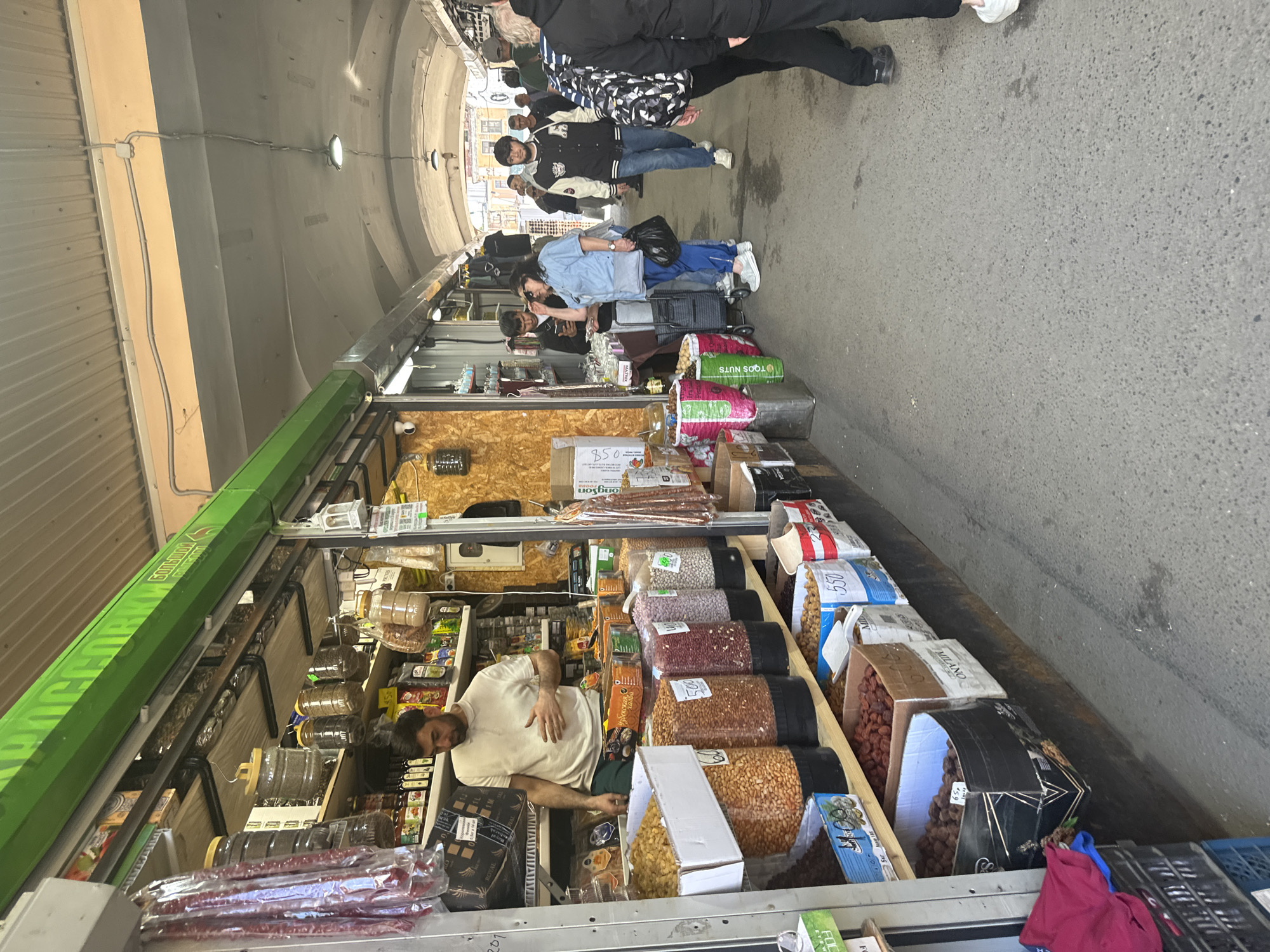 A market stall is depicted in Saint Petersburg, Russia, likely within a covered market or arcade. The stall, painted green along the top, sells various dried goods, nuts, and possibly sausages. A man, presumably the vendor, stands behind the stall, wearing a light-colored polo shirt and dark pants. He is looking down and gesturing with his left hand. In front of him, large clear containers filled with different types of nuts and dried fruits are displayed on shelves. Some containers have handwritten price tags. Behind the vendor, shelves are stocked with packaged goods, jars, and what appear to be dried sausages hanging. To the right of the stall, people are walking along a paved walkway. One woman in a blue shirt and black pants is examining something in her hand. Other individuals, including young men in casual clothing, are also seen walking through the market. The environment appears to be an outdoor or semi-outdoor covered market with overhead lighting.