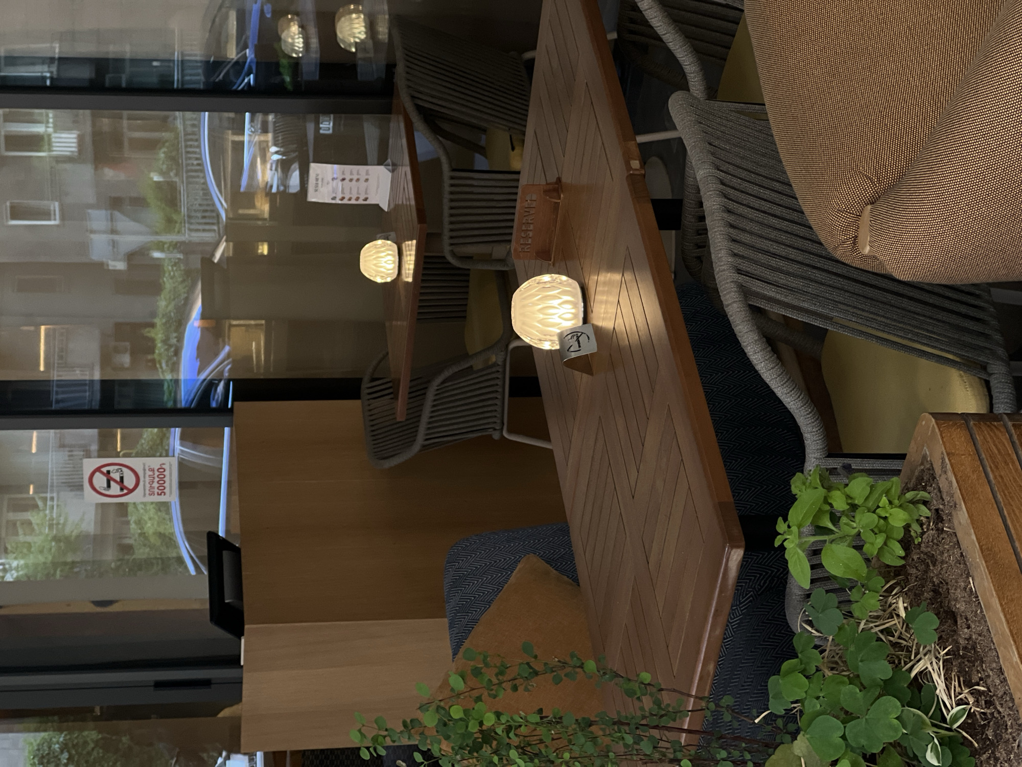 This image depicts a restaurant setting in the city of Unknown, Unknown. It features wooden tables, chairs with gray rope-like backing, and yellow cushions. Some of the tables have small, decorative lamps on them. A window reflects an exterior view of buildings. One window displays a no-smoking sign in a foreign language with the symbol of a cigarette crossed out inside a red circle. There's a potted plant with green foliage visible. The scene appears unoccupied and set for service.