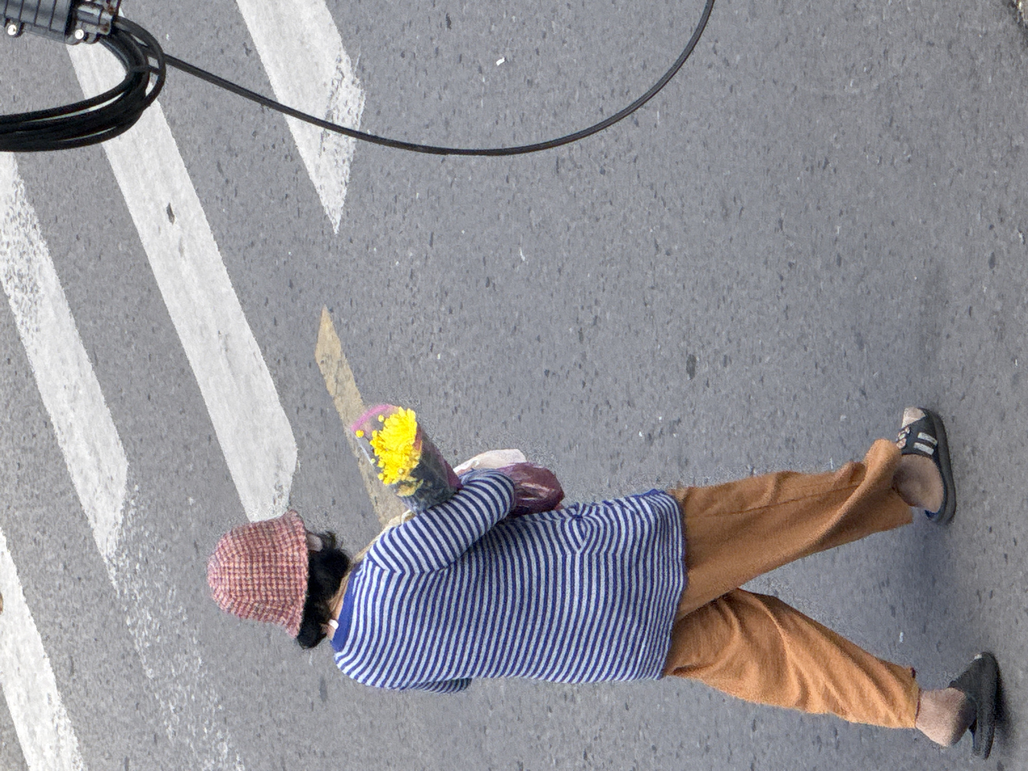 In Da Lat, Vietnam, a person wearing a red and white checkered bucket hat, a blue and white striped long-sleeved shirt, and loose orange pants walks across a street marked with white and yellow pedestrian crossing lines. The individual carries a bouquet of yellow flowers wrapped in clear plastic and a pinkish-purple bag. They are wearing black slip-on sandals. The asphalt street is grey, and to the right of the frame, a thick black cable is visible, attached to a pole or structure. The scene appears to be captured from an elevated perspective, looking down on the pedestrian.