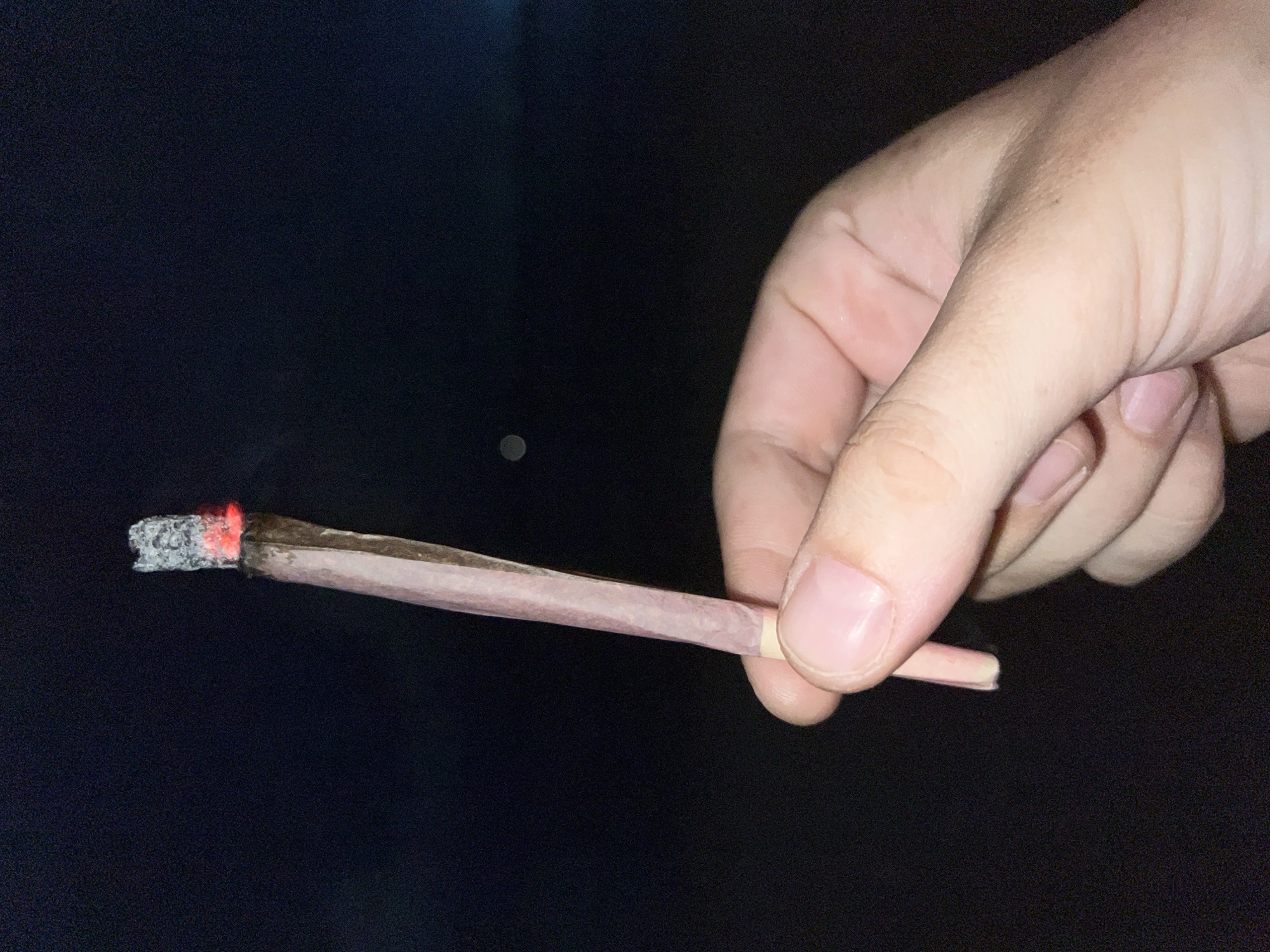 A person's right hand is holding a lit, hand-rolled cigarette (presumably containing marijuana) against a dark background, likely outdoors at night. The cigarette is burning at the tip, displaying glowing embers and ash. The person's hand is in focus, showing skin texture and nails. The background is blurry and completely black. There is no indication of a city location.