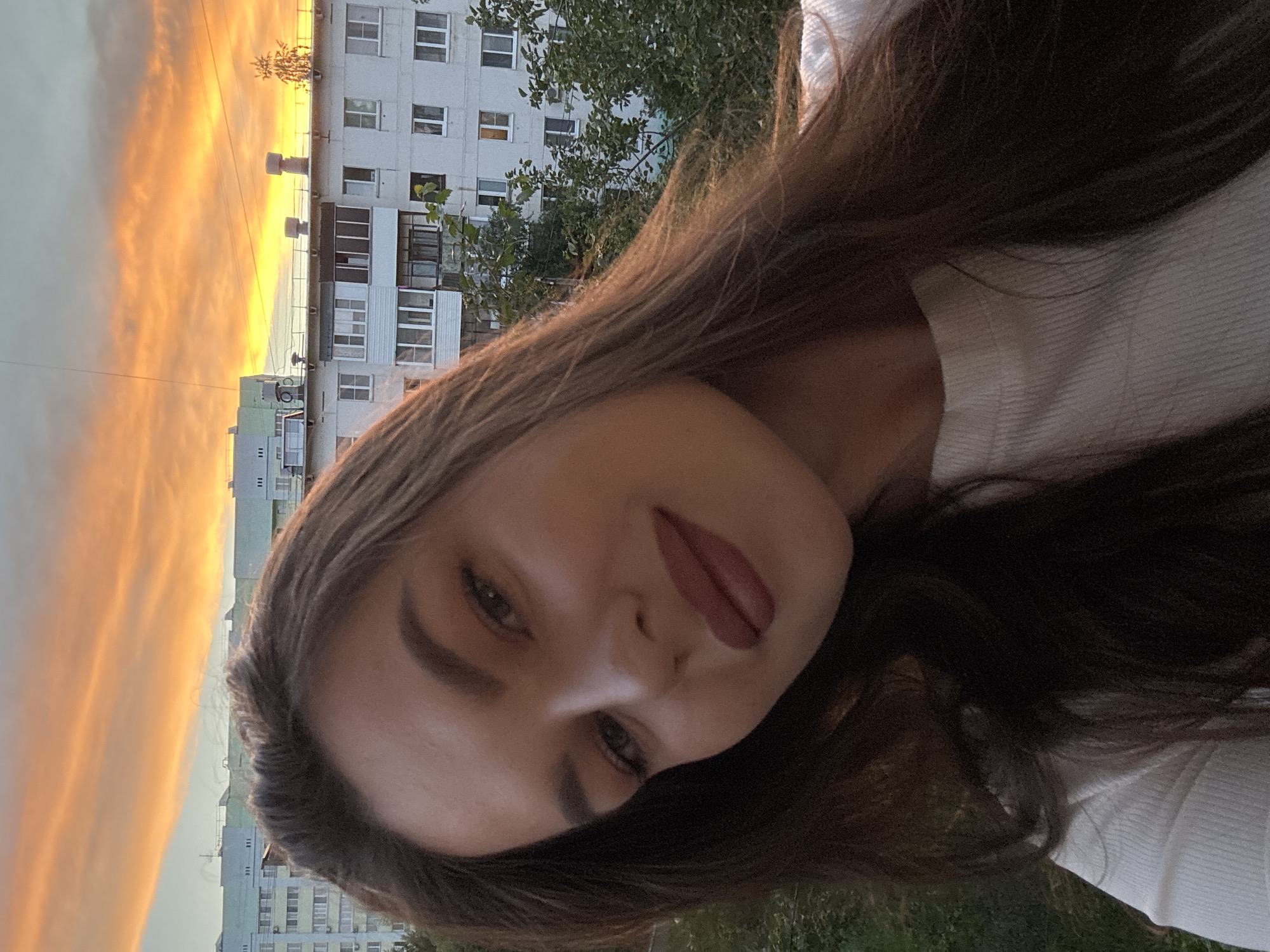 This close-up selfie shows a young woman, with the image rotated 90 degrees clockwise, giving the appearance of her head being tilted to the left. She has long, dark brown hair and is wearing a white ribbed top. Her lips are adorned with dark pink or plum lipstick, and her tongue is slightly visible, conveying a playful or confident expression as she looks directly at the camera.
The background features a vibrant sunset or sunrise, with the sky ablaze in striking orange and yellow hues, transitioning to a lighter grey-blue. Below the colorful sky, several multi-story residential buildings are visible, characterized by multiple windows and balconies, typical of apartment complexes in an urban or suburban setting. Green trees and foliage are scattered among the buildings and in the foreground, suggesting an outdoor environment or a view from a balcony. Power lines or similar overhead wires can be seen stretching across the sky.
The warm, soft light from the setting or rising sun illuminates parts of the woman's hair and face, casting a gentle glow. The architecture of the buildings is consistent with residential areas in Chaikovsky, Russia. The woman appears relaxed, and the overall mood is tranquil due to the sunset lighting. No visible text is present in the image.