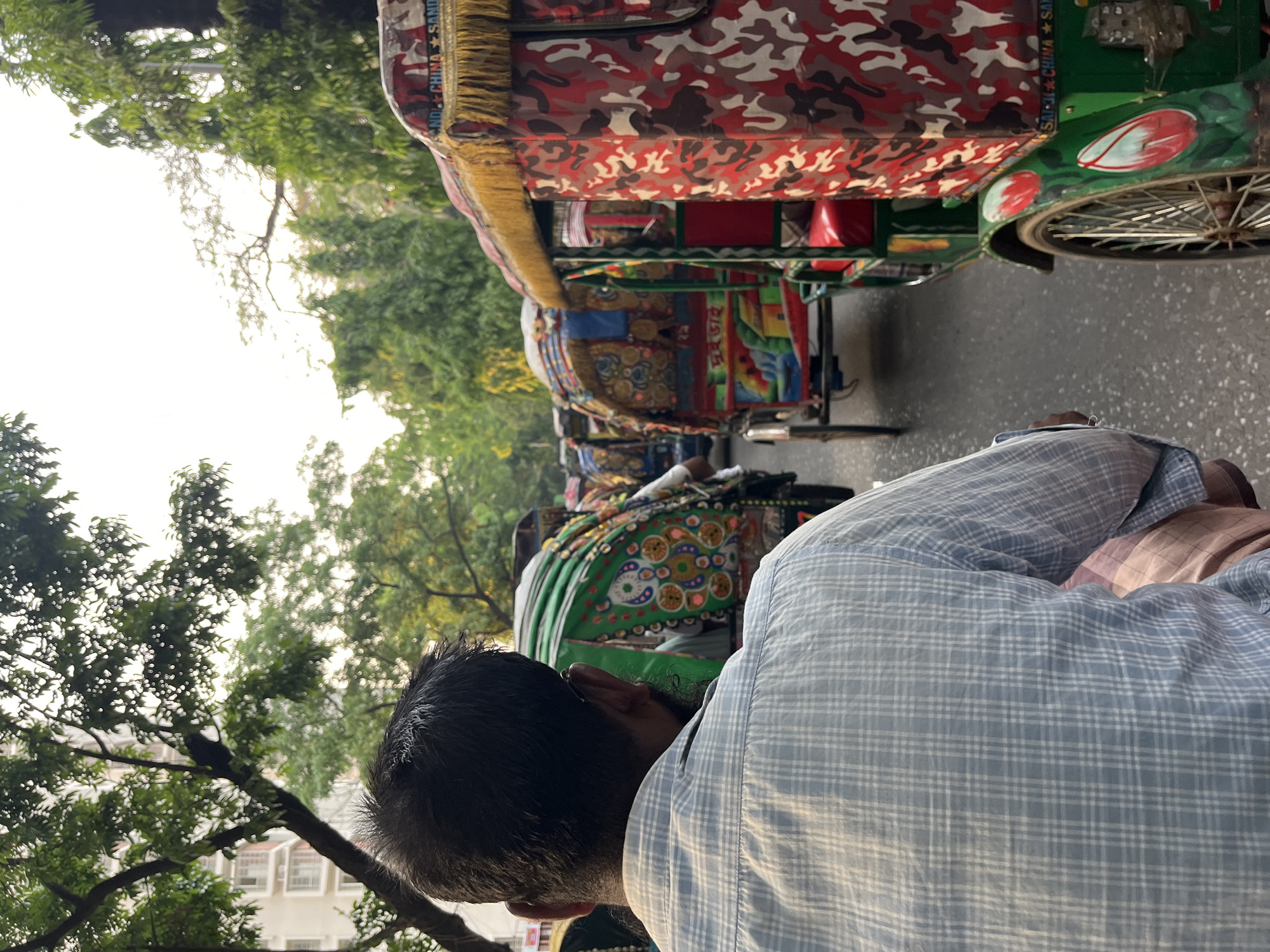 The image shows a row of cycle rickshaws on a street in Dhaka, Bangladesh. The rickshaws are colorful and decorated. A man in a light blue plaid shirt appears to be riding or standing near one of the rickshaws, occupying a portion of the foreground. Trees line the street. The sky is overcast. The perspective is slightly tilted.