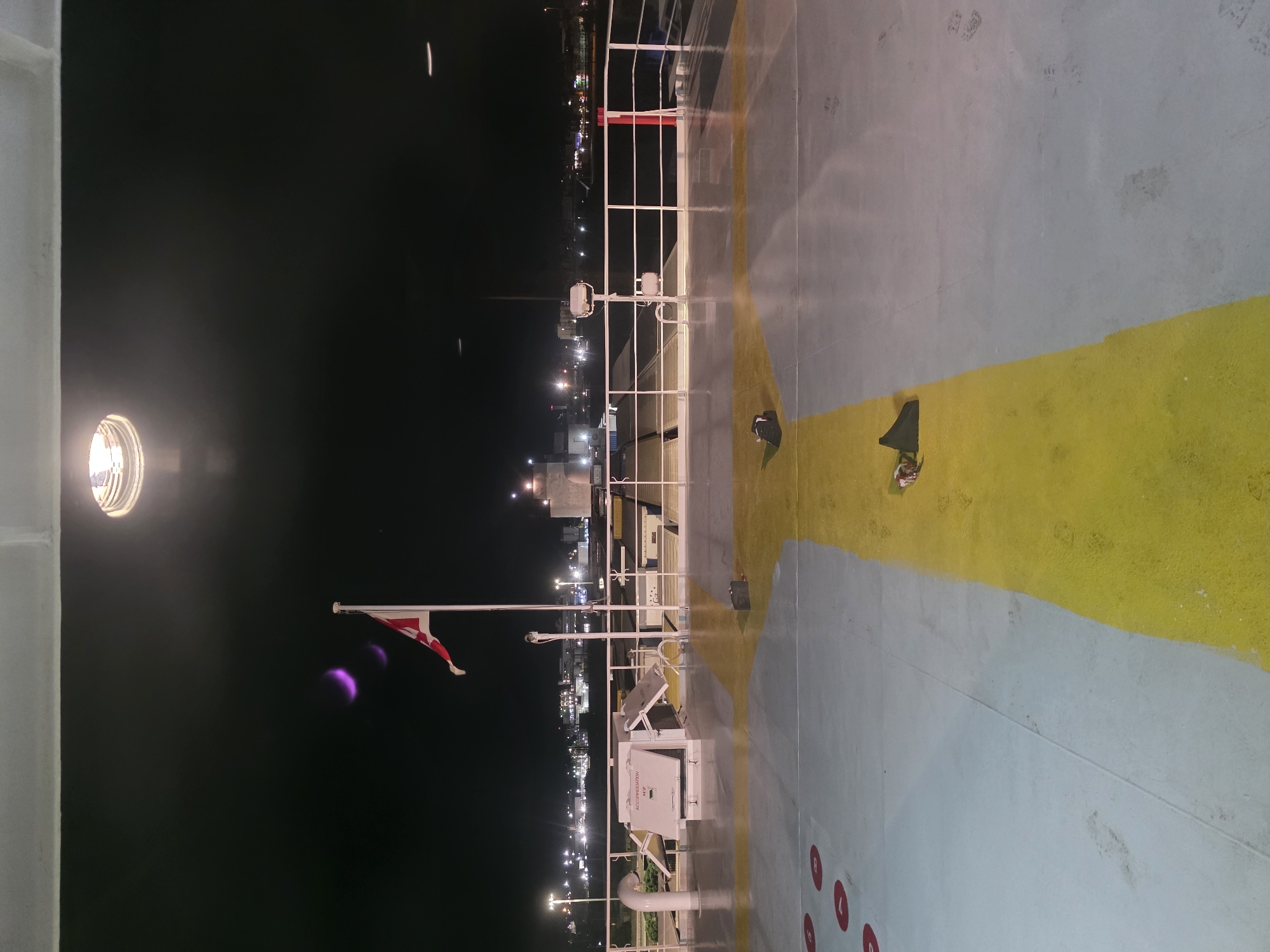 The image depicts the deck of a ferry at night, likely in Brindisi, Italy, given the presence of the Italian flag and the industrial or port landscape in the background.
**Foreground:** The deck is paved and appears wet, with a prominent yellow stripe running diagonally across the scene, bordered by a lighter grey section. Several red circular markings are visible on the grey section, possibly indicating parking spots or safety zones. Two dark, triangular objects, possibly folded tarps or covers, are placed on the yellow stripe. A small black bag is situated near the right edge of the yellow stripe. Faint footprints are visible on the grey deck surface.
**Midground:** A white railing runs along the edge of the deck. Behind the railing, there is a structure with white panels, one of which is labeled "ACCOMMODATION" with an arrow pointing down. Foldable white panels are open on this structure, revealing what appears to be access to an interior. A flagpole with a Danish flag (red with a white cross) is prominently displayed on the right side of the midground, although its placement suggests it might be decorative or a cultural reference rather than signifying Brindisi's nationality. Two floodlights are mounted on poles behind the railing. Further to the right, a red barrier is visible.
**Background:** The background reveals a distant cityscape illuminated by numerous lights. This suggests an urban area or a busy port. Industrial structures and buildings are discernible, along with streetlights and possibly harbor lights. The sky is dark, indicating nighttime, with a bright overhead light source visible at the very top of the image, casting a glare. Faint purple or pink smudges, likely lens flares or light artifacts, are present in the upper left portion of the sky.
**Overall Scene:** The image captures a scene of a ferry preparing for or docked at night. The atmosphere is functional and somewhat industrial, with the lights of the city providing a contrast to the darkness of the sea and sky. The scattered items on deck suggest a temporary stop or a space that is in use.