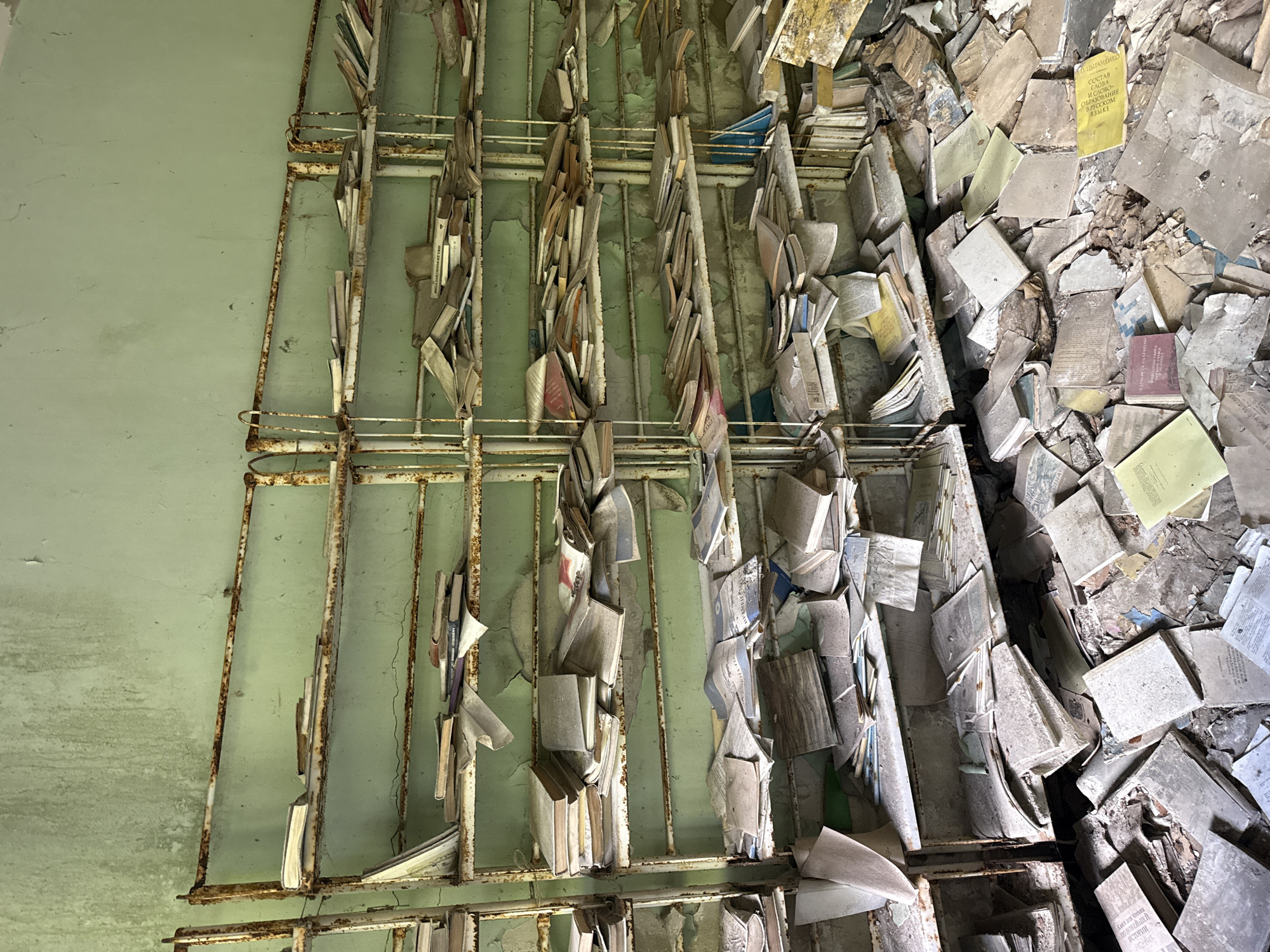 The image depicts a scene within an abandoned building in Prypiat, Ukraine, likely a library or archive, given the overwhelming presence of books. The setting is one of decay and neglect, with peeling green paint on the walls and rusty metal shelving units. The shelves are precariously laden with books, many of which appear to be old and worn, covered in dust and debris. Some books are neatly arranged, while others are haphazardly piled or have fallen to the floor, creating a chaotic landscape of literature.
The books themselves vary in size and color, with some appearing as thick hardcovers and others as thin paperbacks or pamphlets. Their condition suggests a long period of abandonment, with pages yellowed and covers faded. Among the piles, a few text excerpts are visible. One yellow book in the upper right area has text in Cyrillic that includes the phrase "СОСТАВ СЛОВА" which translates to "Word Composition". Another book, a red hardcover, also has Cyrillic text, with the top line appearing to be "ВЕЛИКАЯ ОТЕЧЕСТВЕННАЯ".
The overall impression is one of profound stillness and a sense of lost knowledge. The lack of human presence and the state of disrepair evoke feelings of melancholy and the passage of time. The lighting suggests daylight, but the conditions inside the building are dim and dusty, contributing to the somber atmosphere. The image captures a poignant moment in the history of Prypiat, a city frozen in time following the Chernobyl disaster.