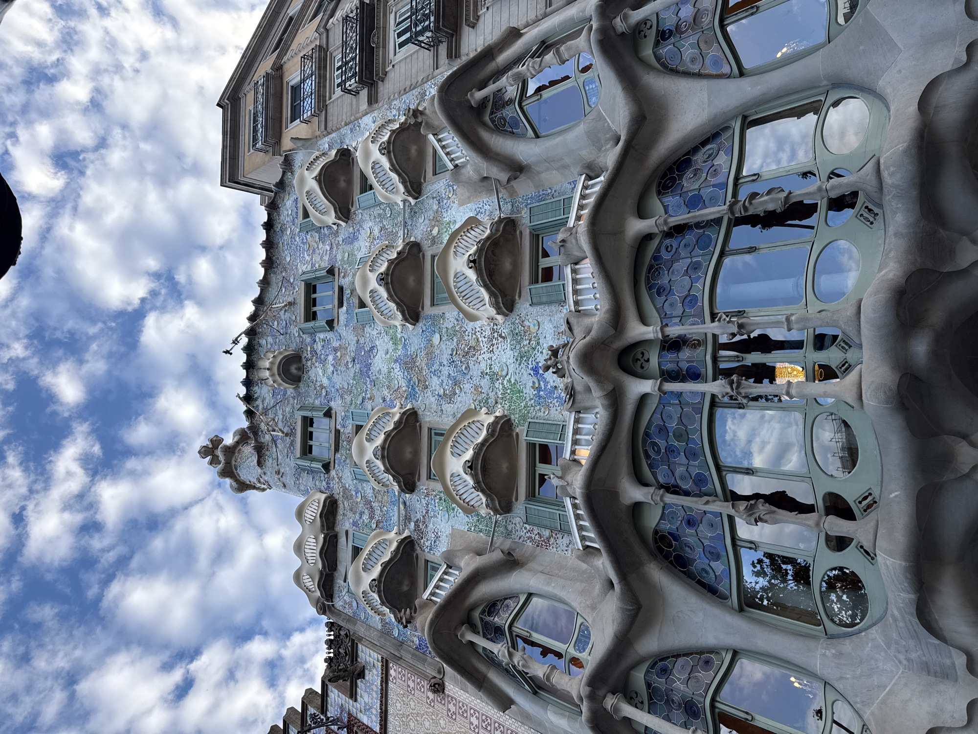 The image shows a section of the exterior facade of Casa Batlló, a building in Barcelona, Spain. The building's facade is characterized by its modernist, organic style, featuring undulating lines, stained glass windows, and skeletal balconies. The exterior walls are covered with a mosaic of colorful ceramics. The windows reflect the sky and clouds. The sky itself is visible behind the building, showing scattered clouds.