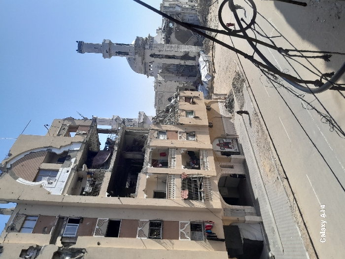 This image presents a wide-angle view of a heavily damaged urban environment in Khan Yunis, Palestinian Territory, under a clear blue sky. The foreground features a dark metal railing, casting prominent shadows onto a paved two-lane road with a dashed white centerline.
To the left, a multi-story residential building, appearing light brown, exhibits extensive structural damage. Multiple floors have external walls collapsed or blown out, exposing interiors filled with debris. Balconies remain on several levels, with articles of clothing, including red and dark garments, visible hanging on a lower-level balcony. An arched architectural detail is discernible on the upper facade. At street level, a dark tent-like structure and a partially visible poster are present.
Across the street, beyond a stretch of unpaved, dusty ground littered with rubble and white tarp-covered areas, the skeletal remains of a mosque are visible. Its tall, light-colored minaret, partially damaged at its apex, stands alongside a ruined dome and other collapsed structures.
Three human figures are present: one individual, clad in dark clothing, walks on the paved sidewalk adjacent to the damaged building on the left. Two smaller figures, also in dark attire, are visible further down the street on the right, near accumulated rubble. The scene is illuminated by bright daylight, casting distinct shadows.