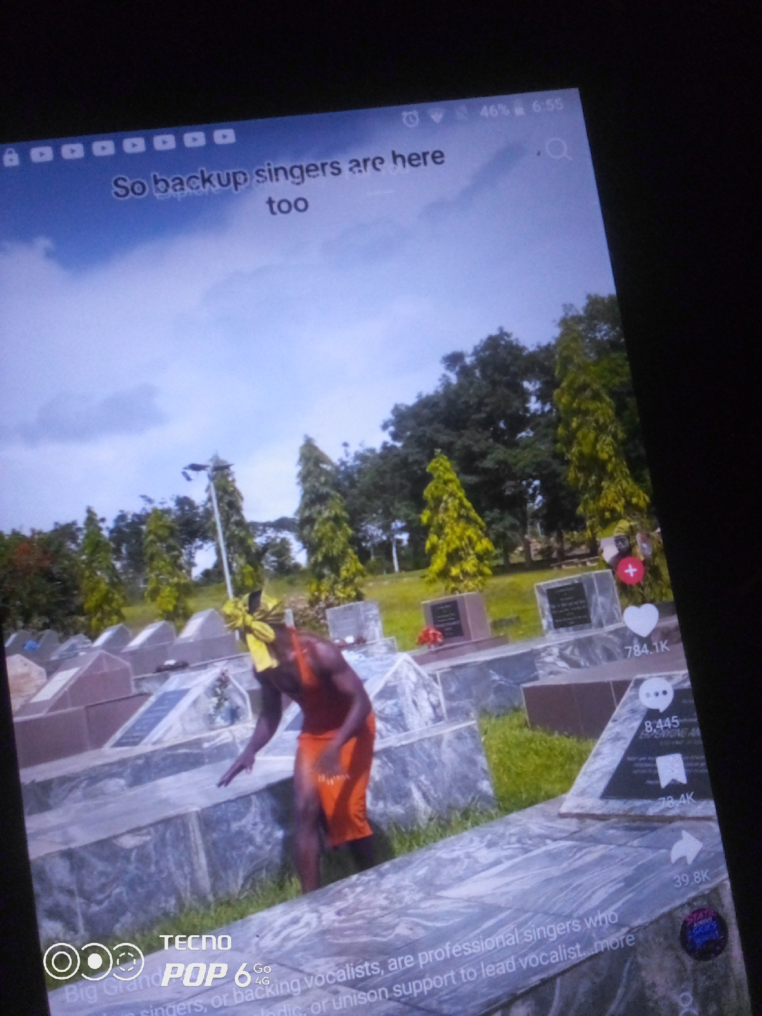The image displays a cellphone screen showing a video with overlaid text. The video depicts a person in an orange garment and a yellow headdress standing in what appears to be a cemetery, with numerous grave markers visible. Trees and a cloudy sky form the background. The screen also shows interface elements like a battery indicator (46%), time (6:55), a search icon, a heart icon with a number (784.1K), and a comment icon with a number (8,445). At the bottom of the screen, a TECNO POP 6 Go logo is visible, along with text that reads "Big Grand singers, or backing vocalists, are professional singers who ...support to lead vocalist...more". The specific location of Howayi, Nigeria, is not visually verifiable from the image content itself.