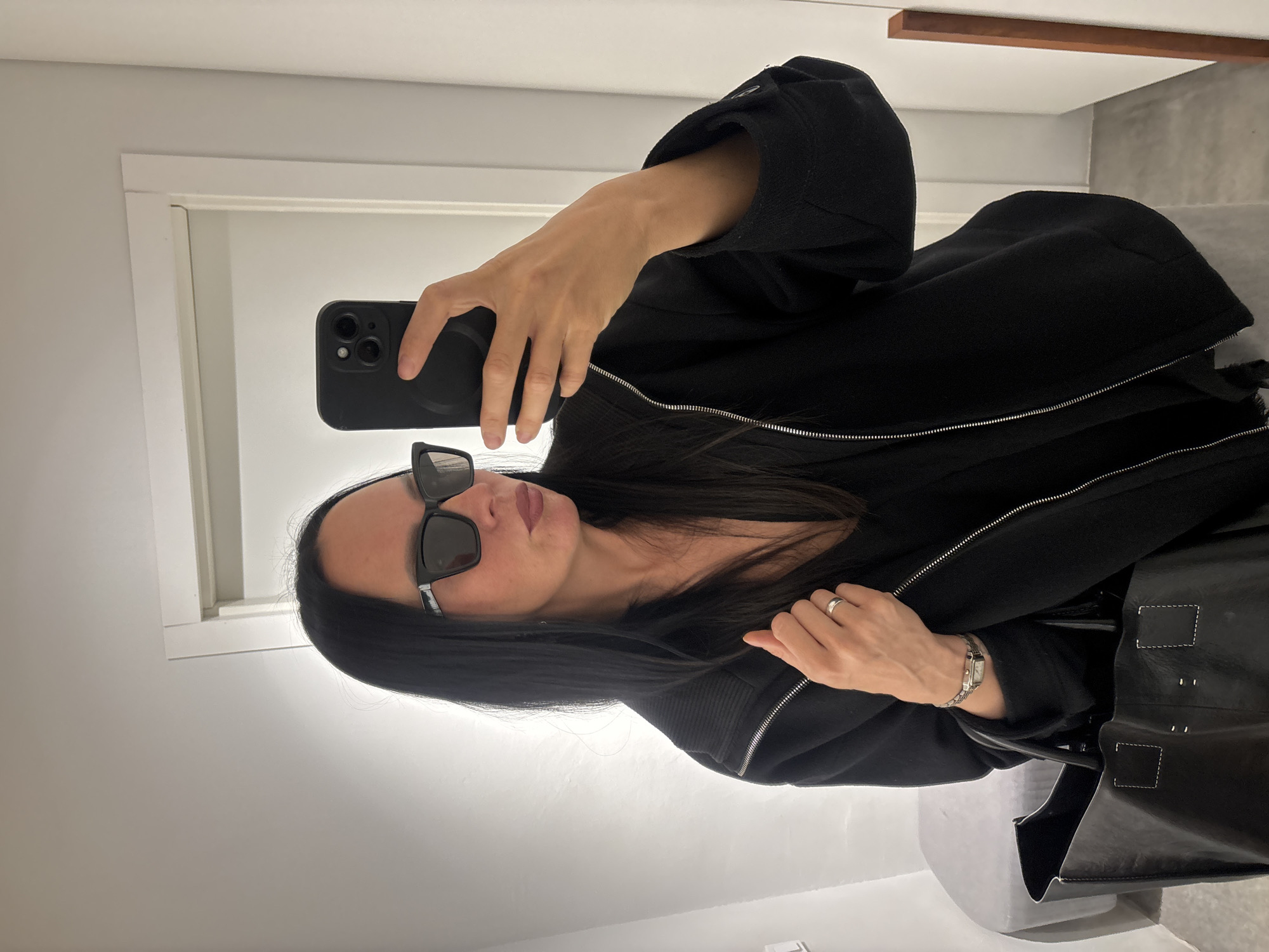 A woman wearing dark sunglasses and a black jacket with a silver zipper is taking a selfie with a black smartphone. The setting appears to be indoors, with white walls and neutral-toned flooring. A portion of a handbag is visible in the lower right. The location is not identifiable as Chaikovsky, Russia, from the image alone.