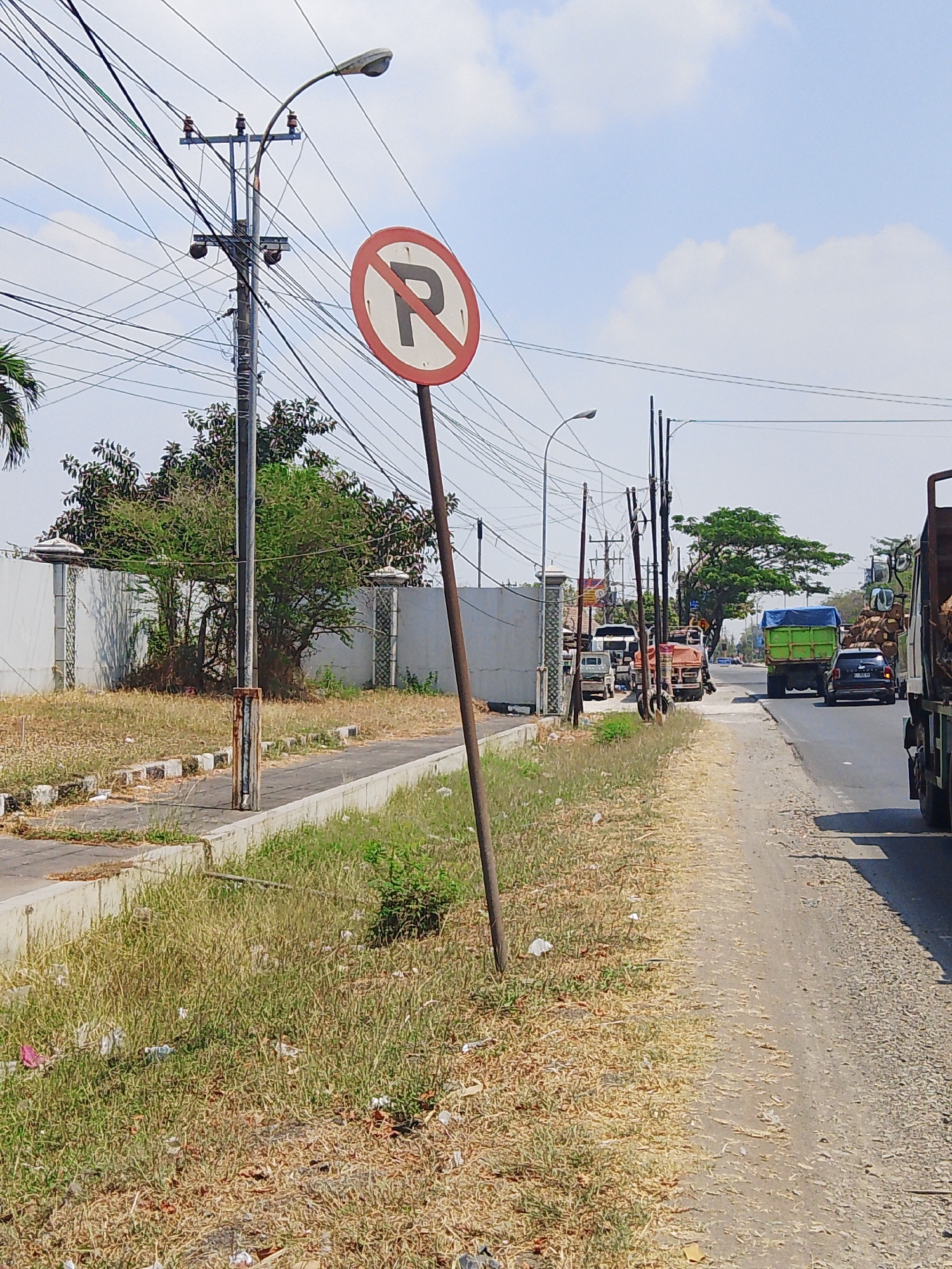 The image shows a street scene in Bolo Krajan, Indonesia. A "no parking" sign is prominently displayed on the left side of the road. The road is flanked by overgrown grass and a concrete barrier. Several vehicles are visible on the road, including trucks and cars. Overhead, a complex network of electrical wires and a streetlamp are present. The sky is blue with some scattered clouds. The overall impression is of a sunny day in a somewhat unkempt urban or semi-urban environment.