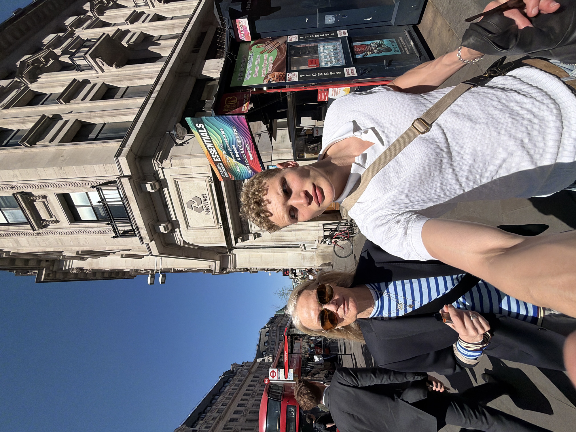 An outdoor selfie captures two people on a sunny street in London, United Kingdom. In the foreground, a young man with curly blonde hair and a white textured shirt holds the camera, looking towards it. Next to him, a woman with blonde hair, sunglasses, a blue and white striped top, and a black jacket also looks at the camera. Her hand, adorned with bracelets, holds a small object. Partially visible in the bottom right is another person in a dark suit.
The setting is a busy city street, flanked by grand, ornate buildings characteristic of London architecture, including one with the "NatWest" logo. To the right of the subjects, a series of kiosks are visible. A prominent sign for "ESSENTIALS" features a rainbow gradient and advertises "Mobile Phone Accessories & Repairs," "Vape Liquids & E-Cigarettes," "Souvenirs," and "Cold Drinks." Other kiosk signs promote "VAPING," "Big Bus Tour Tickets," "Churros Waffles," "Freshly Squeezed Orange Juice," and "Cold Bottled Water," with several "SOLD HERE" notices. Text like "BIG LITTLE ARGYLL STREET KIOSKS" and "YOUR PAPE STORE" (likely "VAPE STORE") are also present.
In the background, a bright blue sky is visible above the buildings. A red double-decker bus, a quintessential London sight, is stopped or moving slowly, bearing the number "159" and what appears to be part of a "HOP-ON HOP-OFF" tour bus sign. The scene is well-lit by bright sunlight, casting distinct shadows, indicating a clear, sunny day, likely morning or early afternoon. The overall impression is one of a lively urban environment.