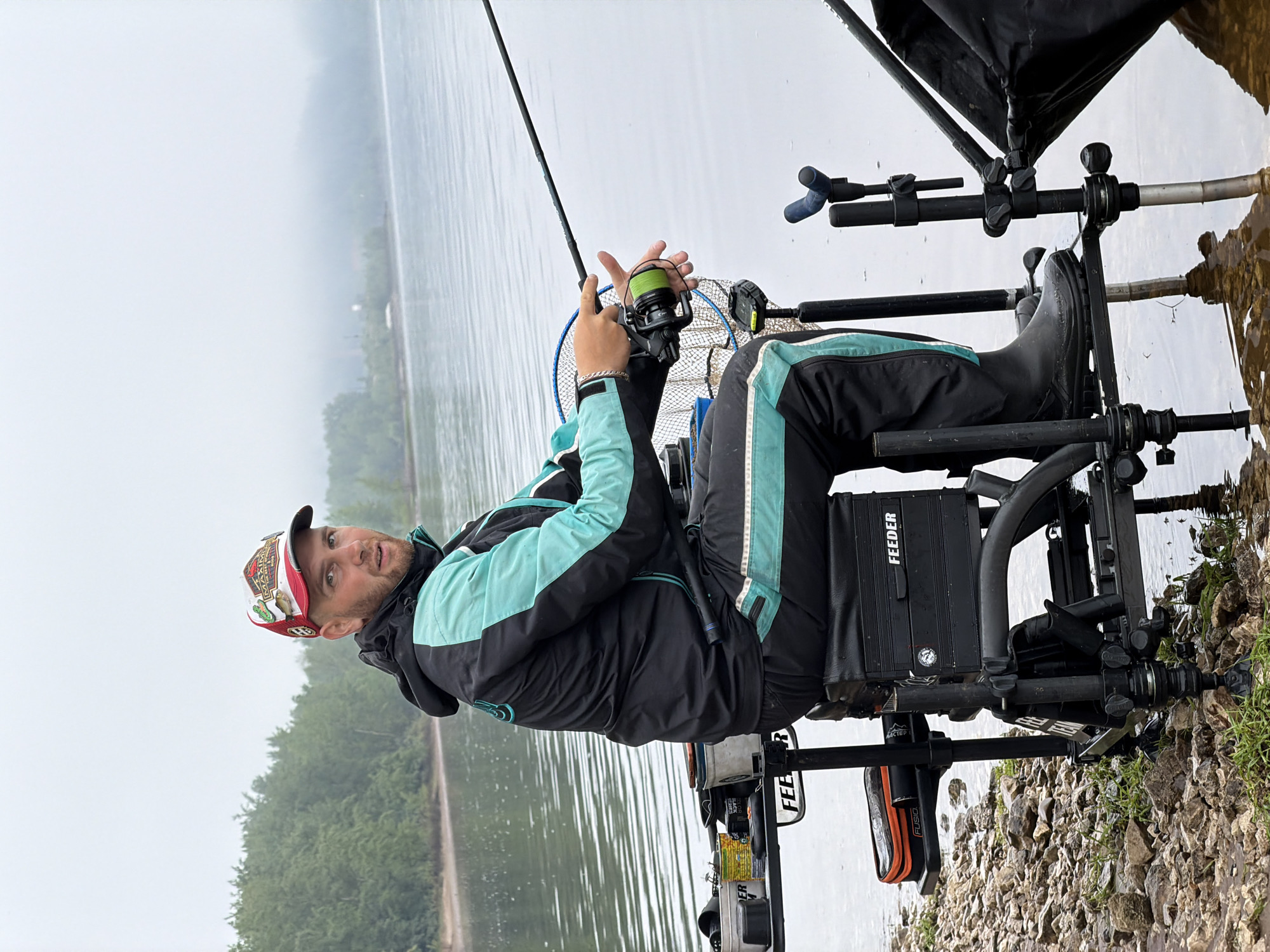 The image captures a male angler engaged in feeder fishing on the rocky bank of a large body of water, likely a river or lake, in what is identified as Chaikovsky, Russia. The man, with a light beard, wears a baseball cap with red and white designs, a black and turquoise hooded jacket, and black pants with turquoise stripes. He is seated in a robust black feeder fishing station, which is partially extended over the water.
He holds a fishing rod with both hands, his right hand positioned on the reel, which is spooled with conspicuous neon green fishing line. His expression is one of calm focus, looking slightly towards the viewer. The fishing station is equipped with various attachments, including a rod rest, a black canopy or umbrella, and a storage unit. The word "FEEDER" is prominently visible in white text on the side of the storage box, and also on another piece of equipment. Additionally, "ACTEC" and "FUSION" are visible on attached gear. The man wears black rubber boots, suggesting preparedness for waterside conditions.
The scene unfolds on an overcast day, indicated by the soft, diffused light and the absence of harsh shadows. The calm water reflects the light sky, extending to a distant, tree-lined bank covered in lush green foliage. The immediate foreground reveals a ground composed of pebbles and small rocks mixed with sparse green grass, where the fishing station's legs are planted, some partially submerged in the water at the edge. The atmosphere is one of tranquil concentration on the activity of fishing.